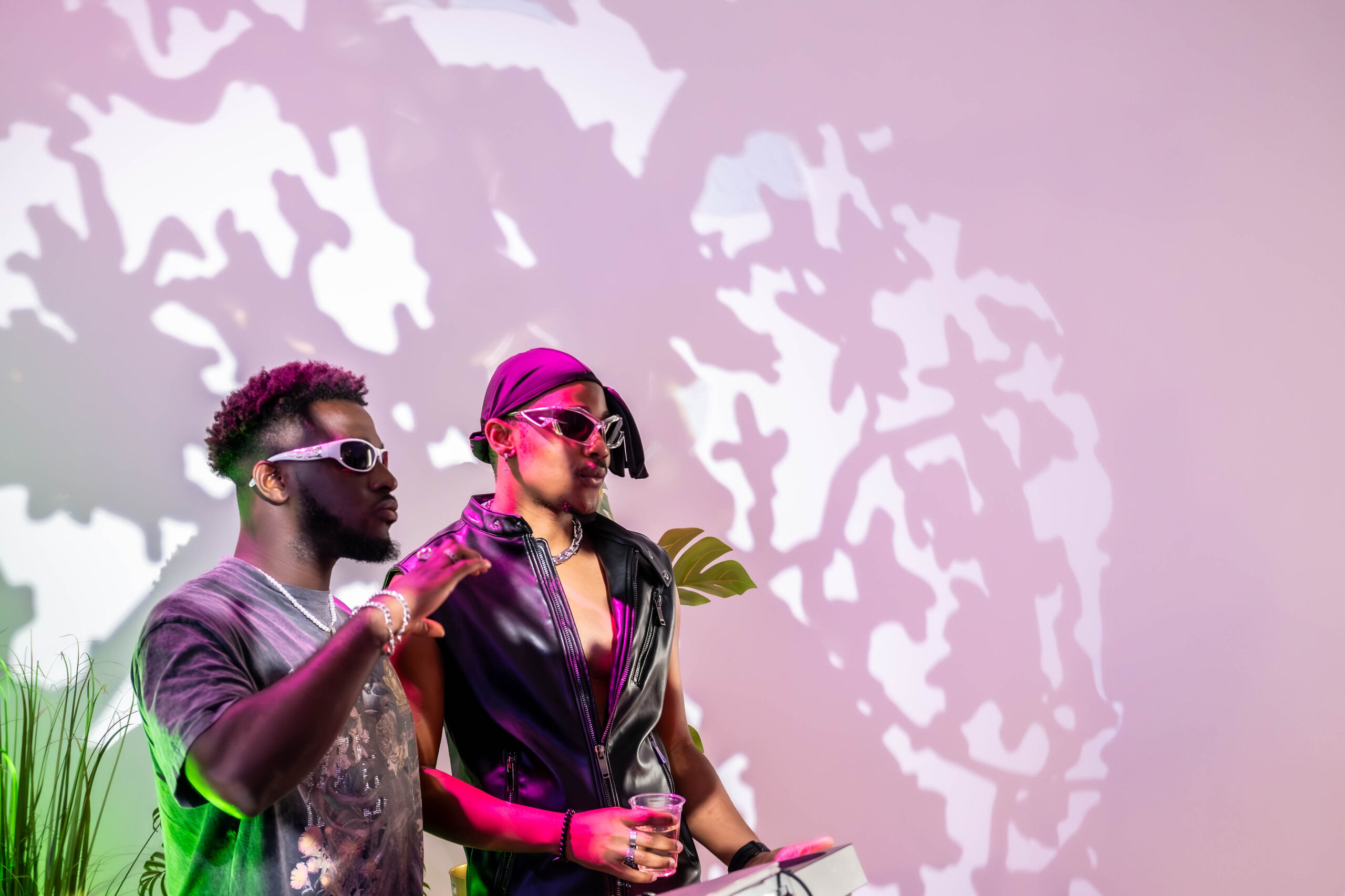 Two stylish men wearing futuristic sunglasses and modern streetwear, illuminated by pink lighting against a wall with organic shadow projections.