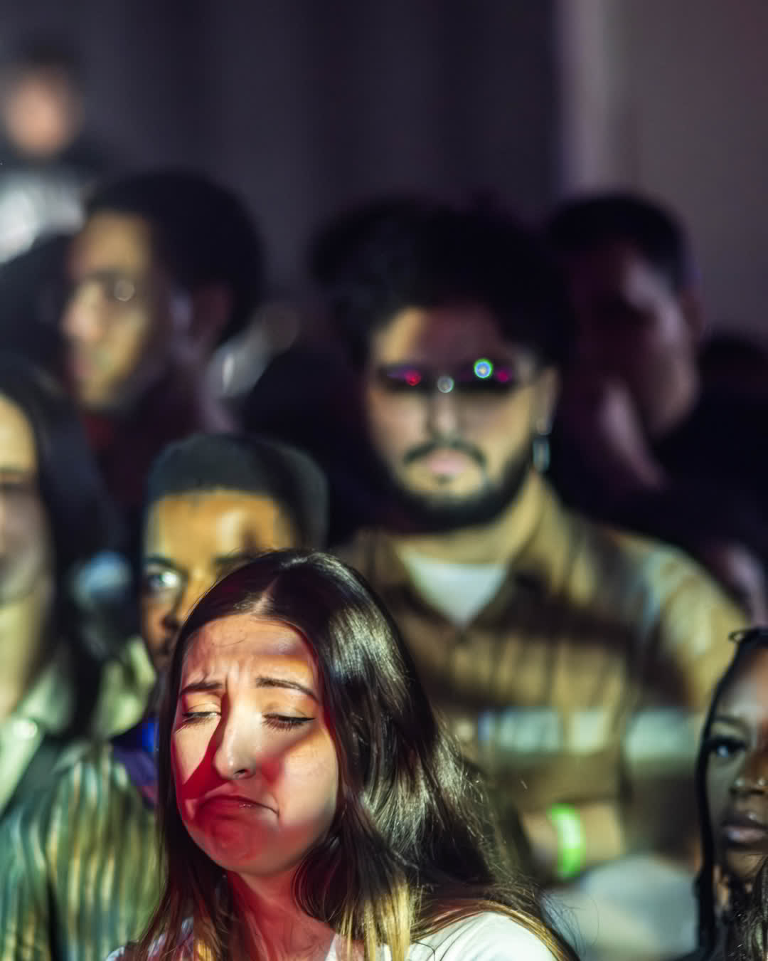 A young woman in a concert crowd with her eyes closed, showing intense emotion, illuminated by red and white stage lights.