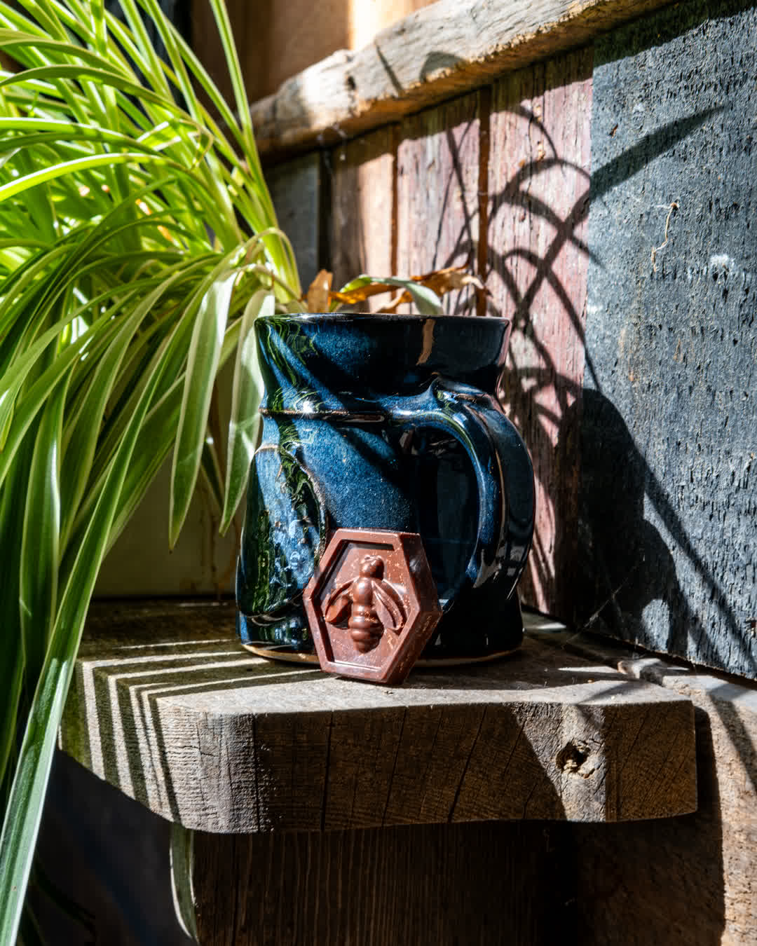A blue, handcrafted ceramic mug sitting on a rustic wooden ledge in harsh sunlight next to a piece of chocolate shaped like a honeycomb with a bee on it.