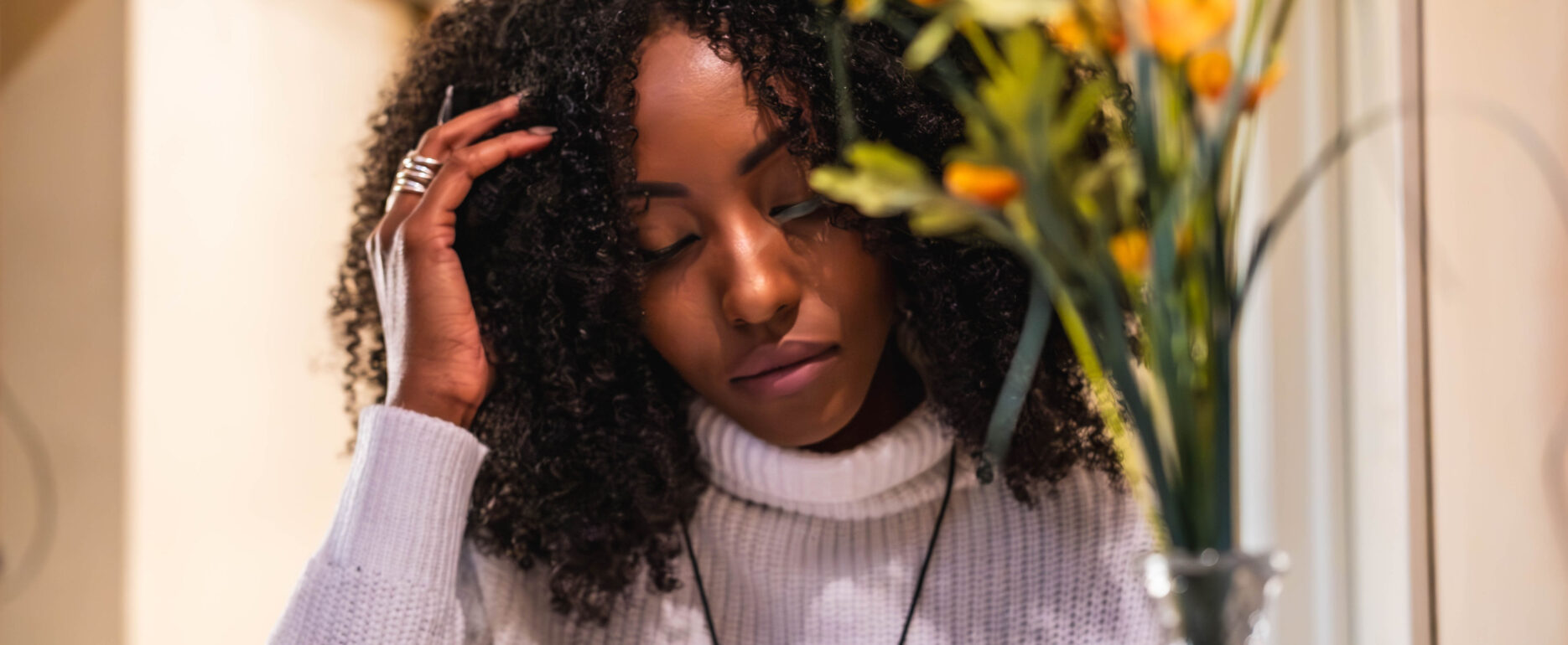 A woman with dark curly hair wearing a white turtleneck, looking down thoughtfully with her hand in her hair, framed by blurred flowers.