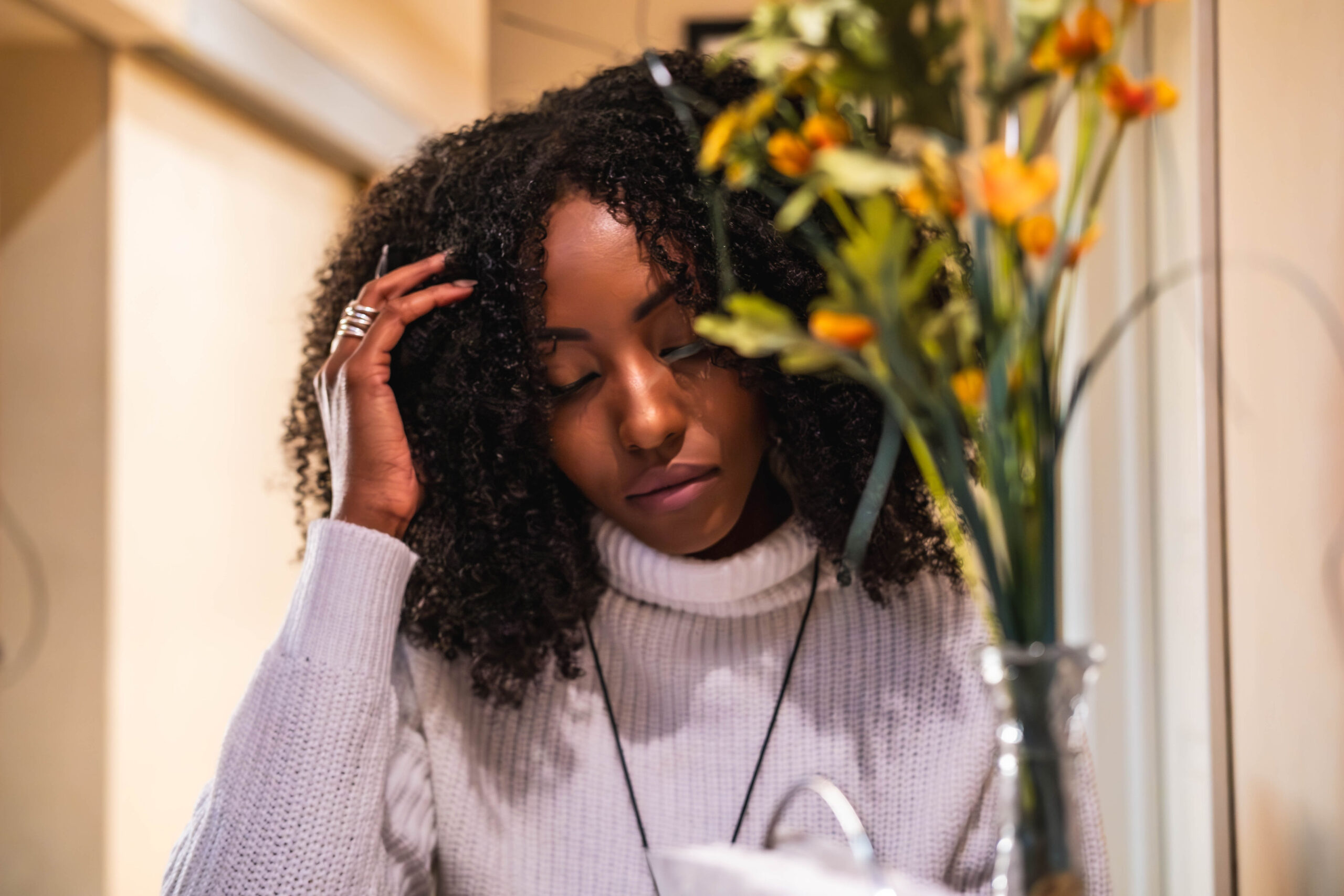A woman with dark curly hair wearing a white turtleneck, looking down thoughtfully with her hand in her hair, framed by blurred flowers.