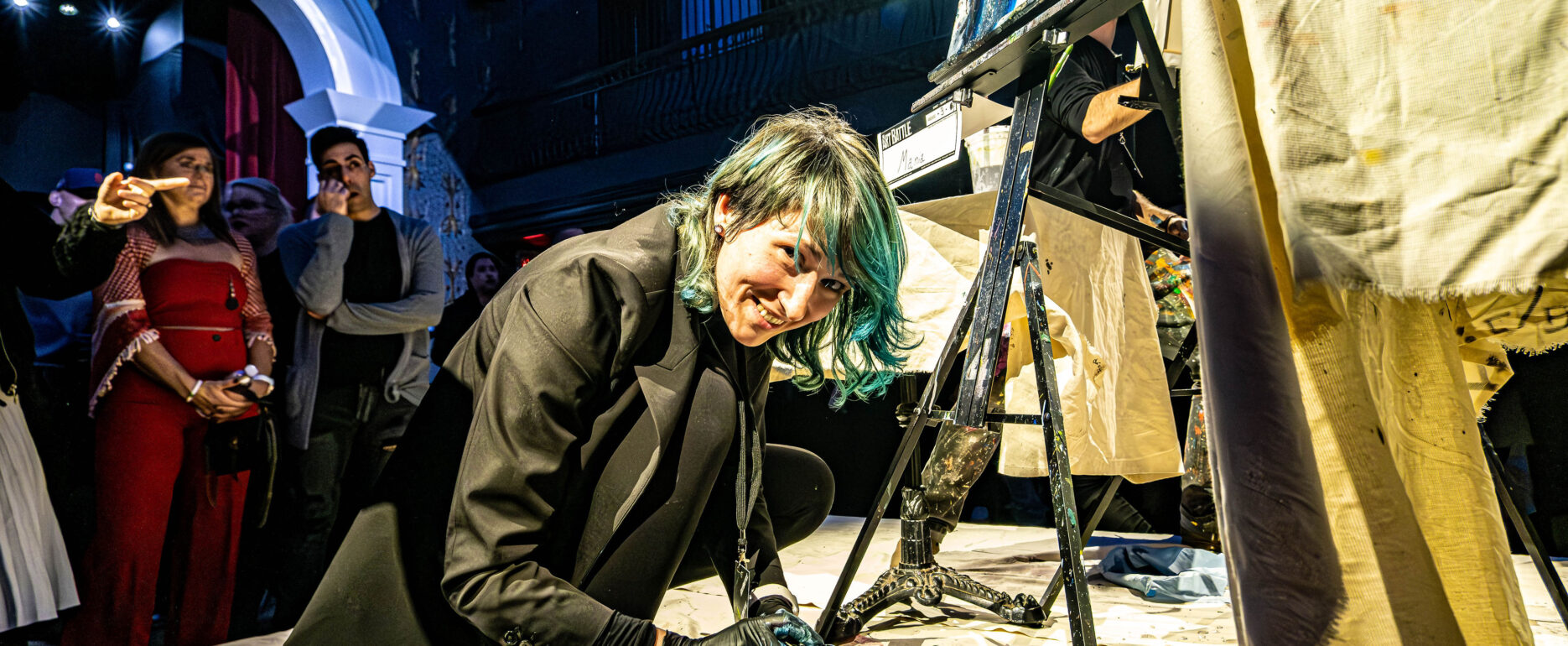An artist with teal hair smiling at the camera while kneeling on a drop cloth to paint live on stage in front of an audience.