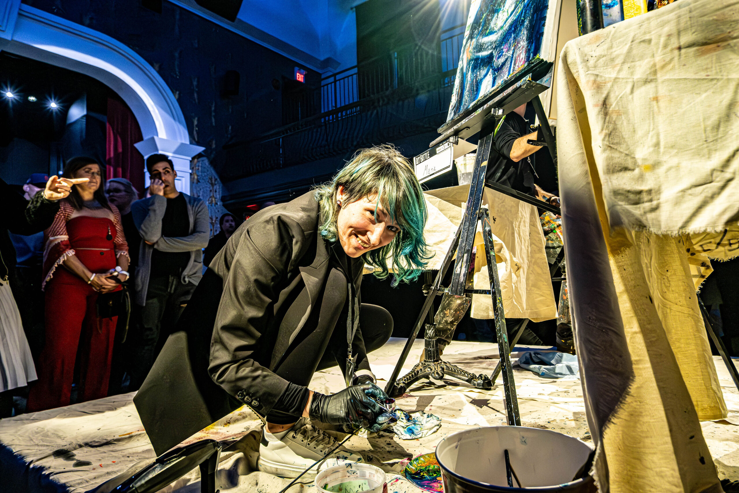 An artist with teal hair smiling at the camera while kneeling on a drop cloth to paint live on stage in front of an audience.