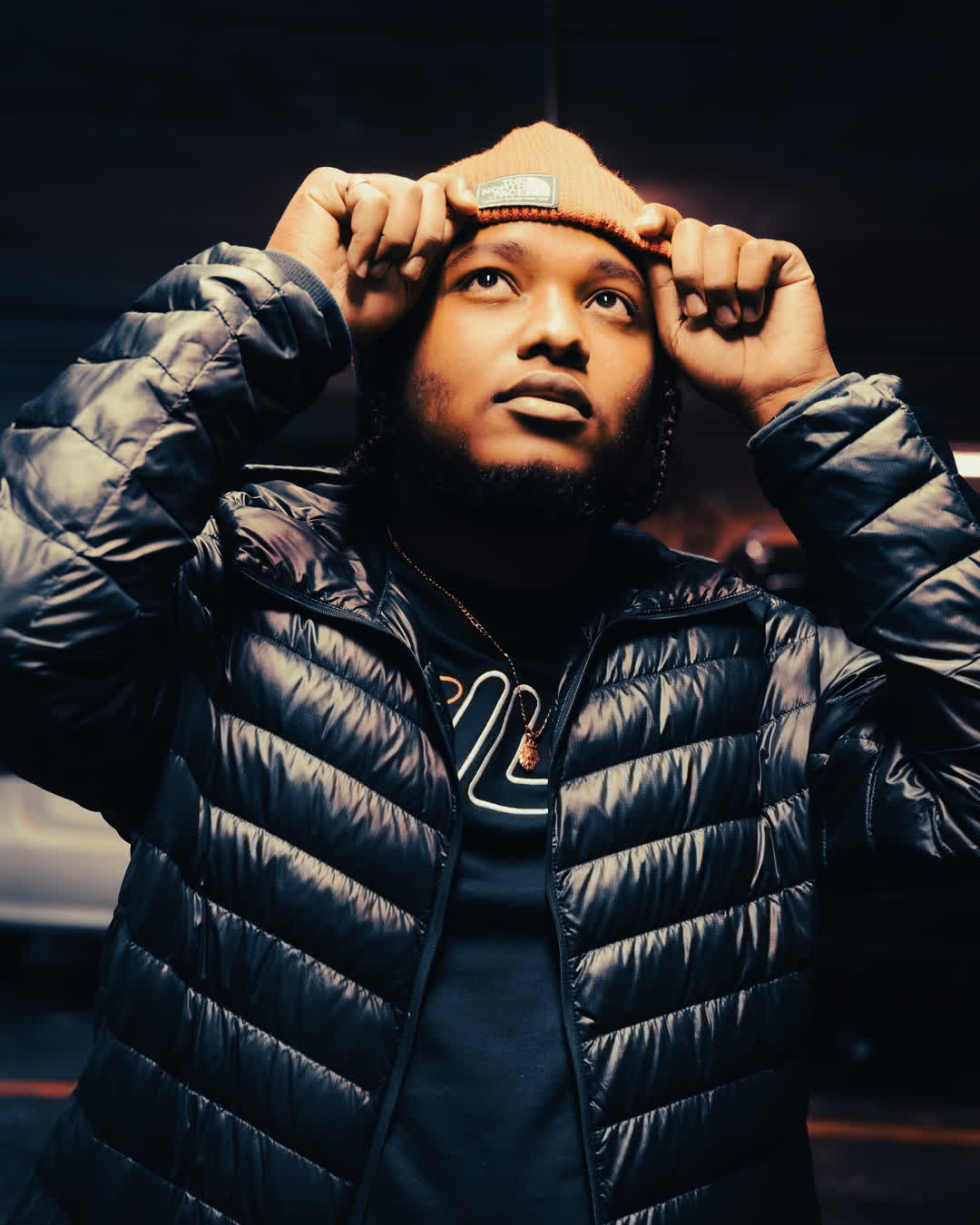 A young man in a black puffer jacket adjusting his orange beanie, looking upward into dramatic, cinematic lighting.