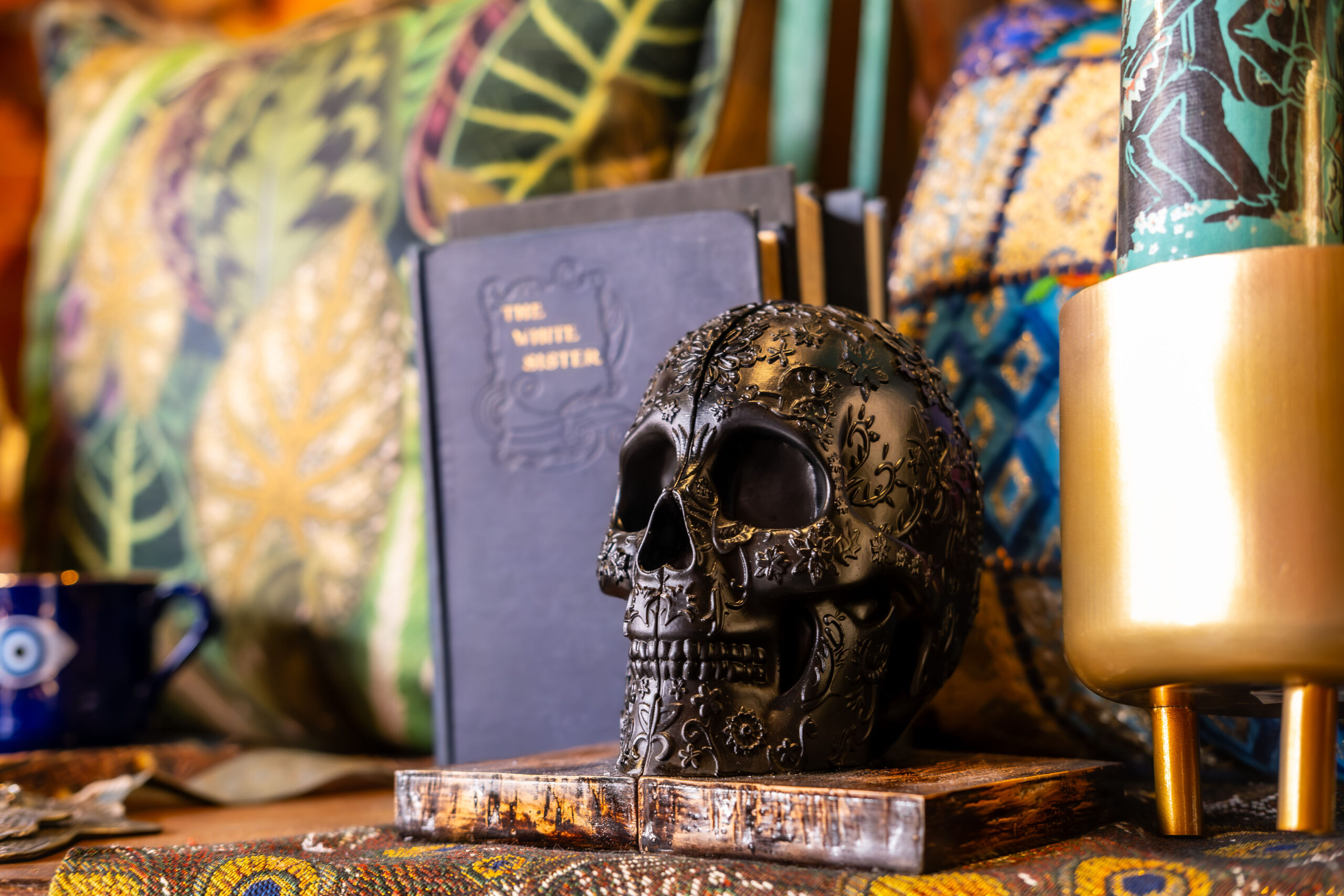 Ornate black carved sugar skull resting on a vintage book next to brass decor and vibrant textured pillows.
