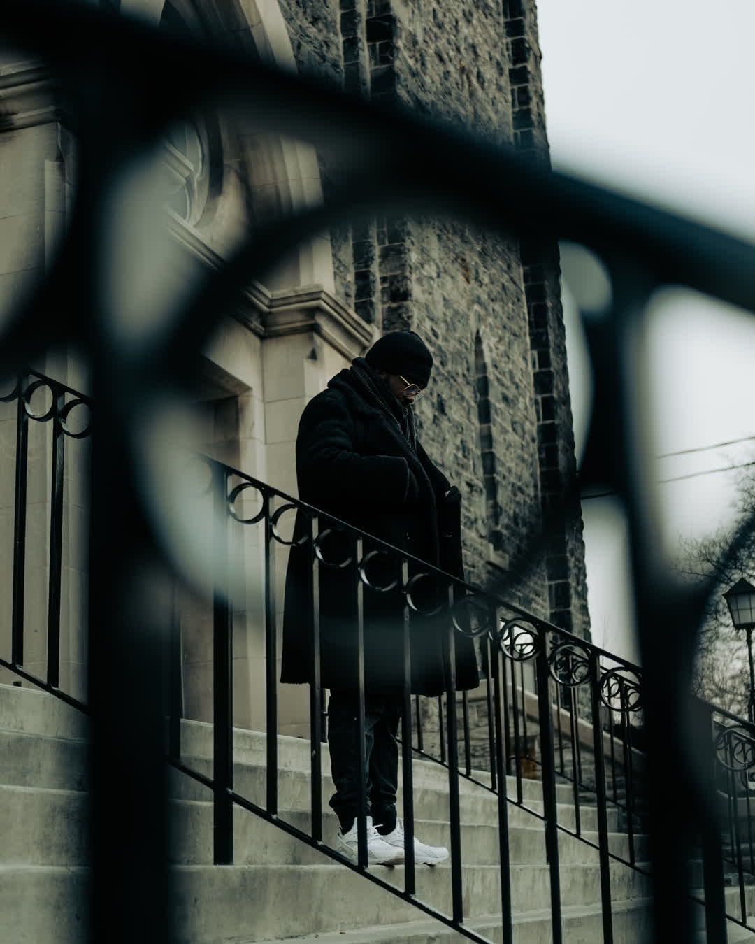 Moody urban street photography of a man in a black coat and beanie standing on stone steps outside a historic church, framed through a dark wrought-iron fence.