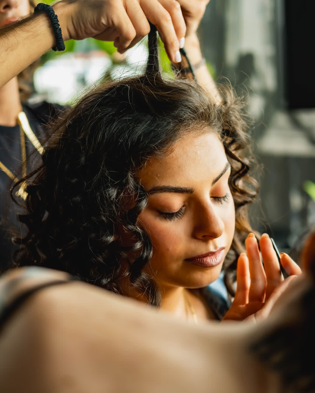 Close-up of a beautiful woman with dark curly hair getting styled by a professional makeup artist and hair stylist in warm, natural lighting.