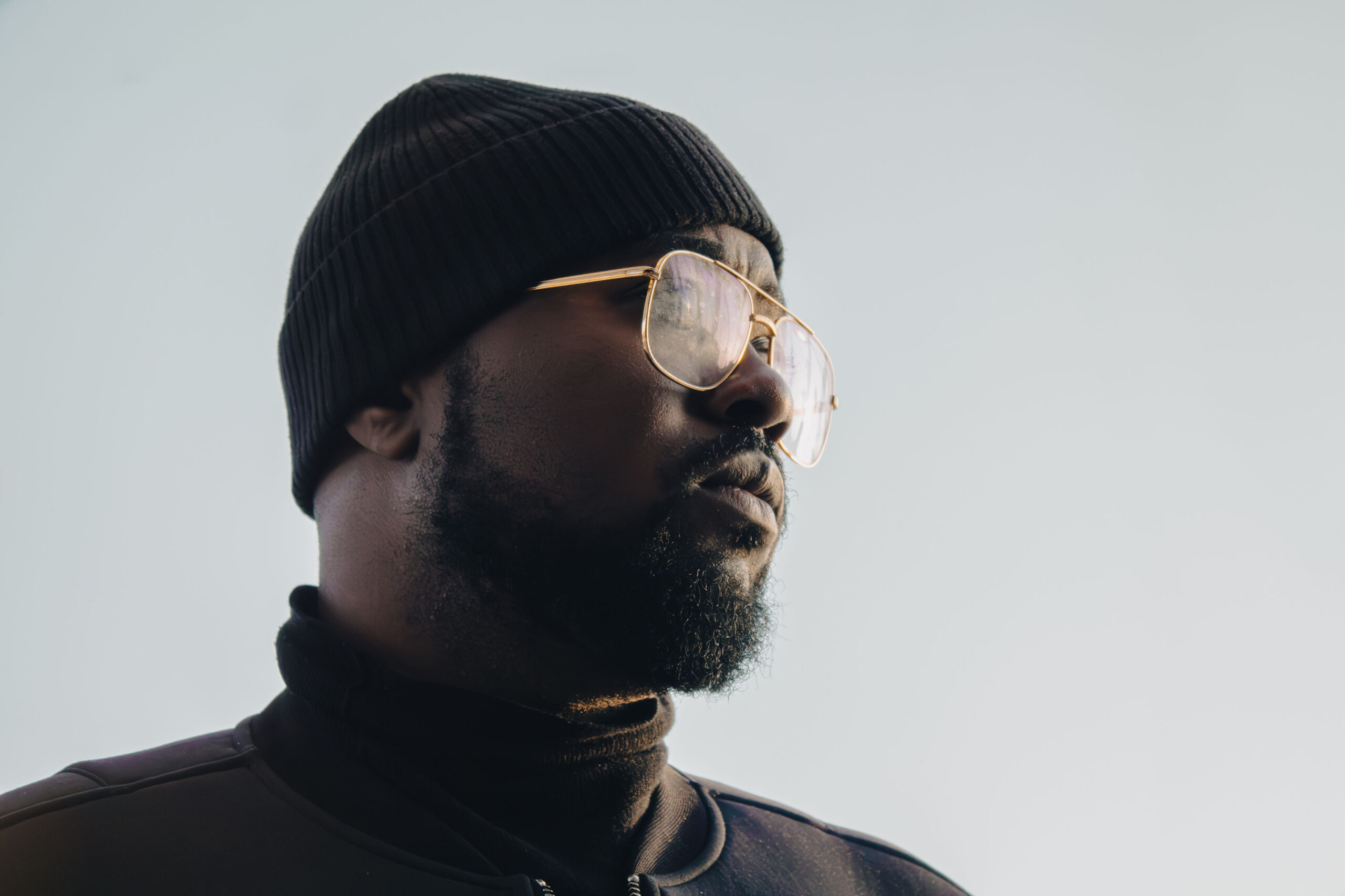 Low angle cinematic portrait of a Black man with a beard wearing a black beanie and gold-rimmed aviator glasses looking confidently into the distance.