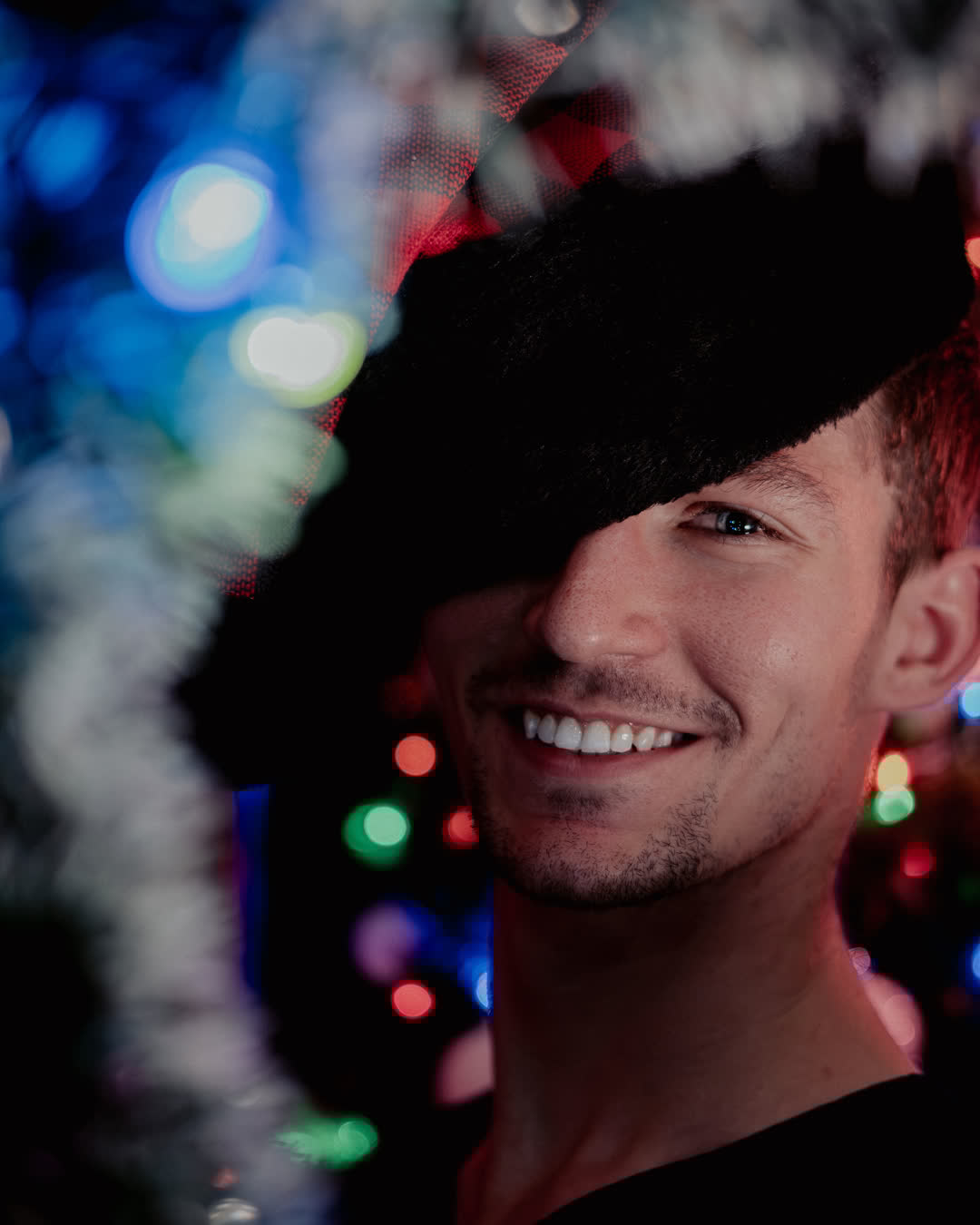 Smiling portrait of a man partially hidden by a black fuzzy hat, surrounded by colorful blurred bokeh fairy lights in the background.