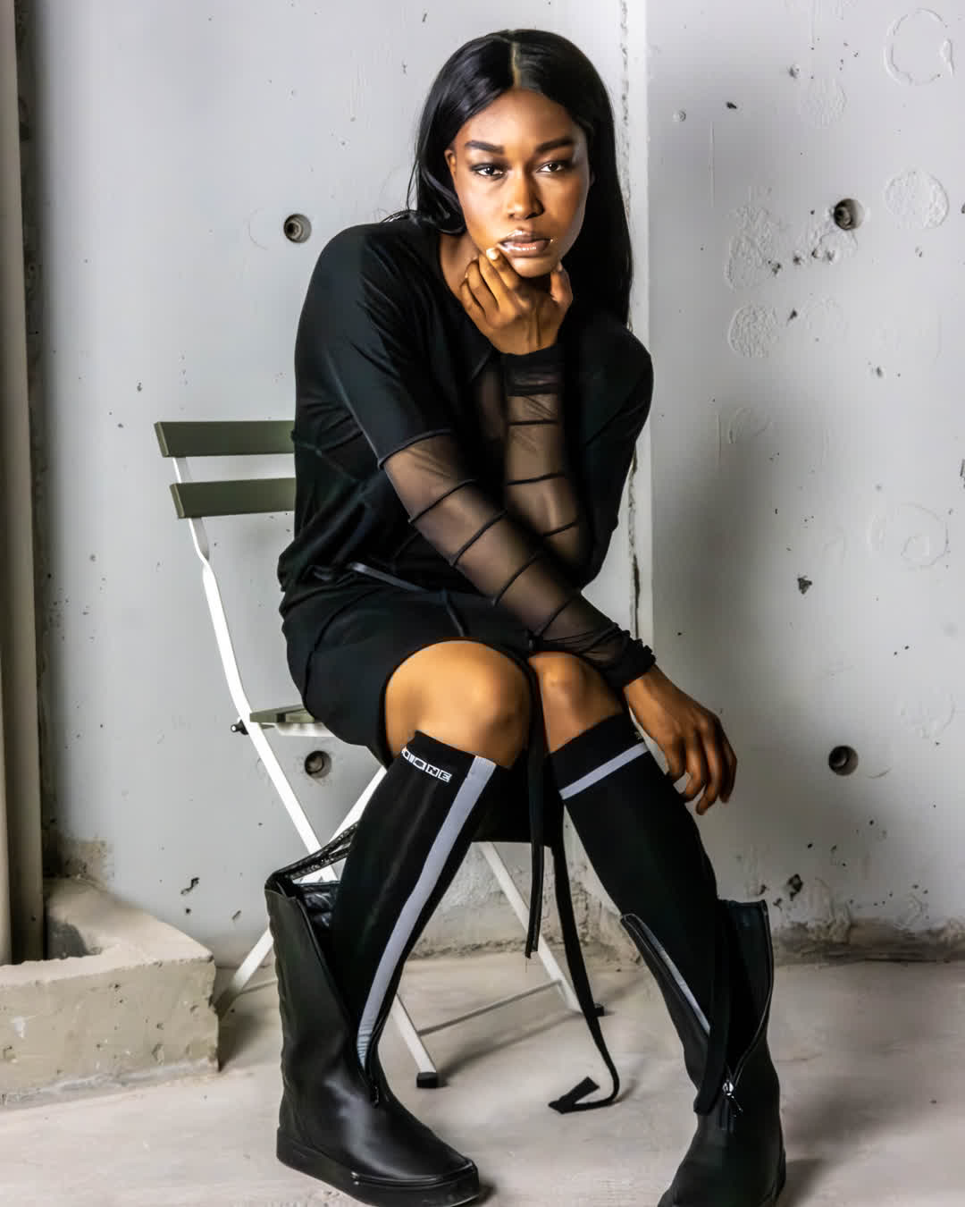 Fashion editorial portrait of a woman in a black dress and striking knee-high boots sitting on a metal chair in an unfinished industrial concrete room.