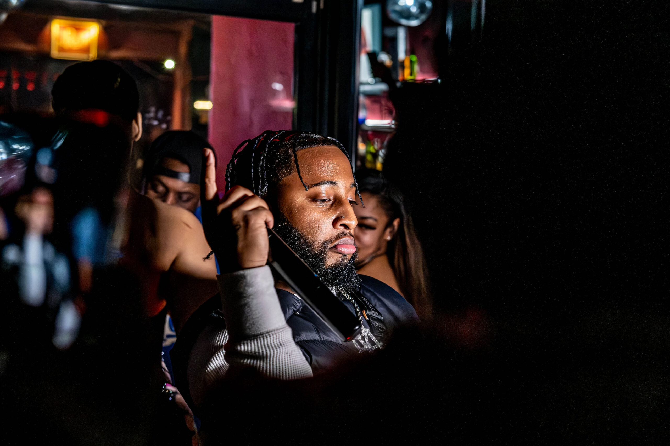 Candid low-light event photography of a man with braided hair holding a phone in a crowded, dimly lit club environment with purple neon lighting.