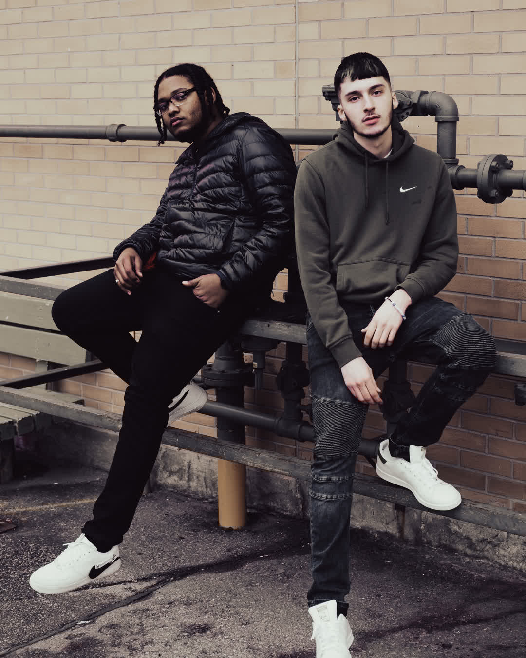 Two young men in streetwear posing casually against an industrial brick wall with metal piping.