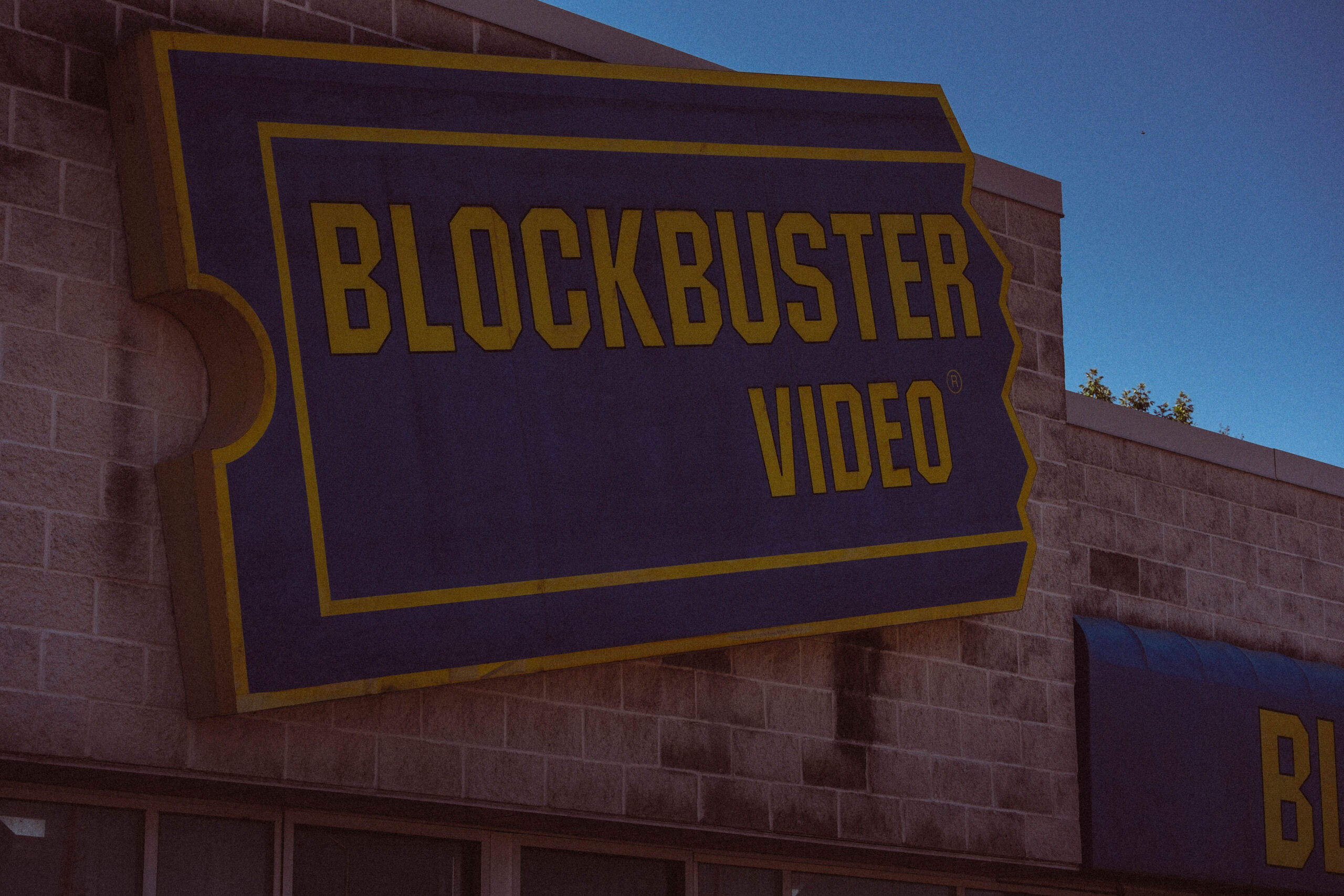 A weathered, glowing Blockbuster Video sign mounted on a brick storefront against a moody twilight sky