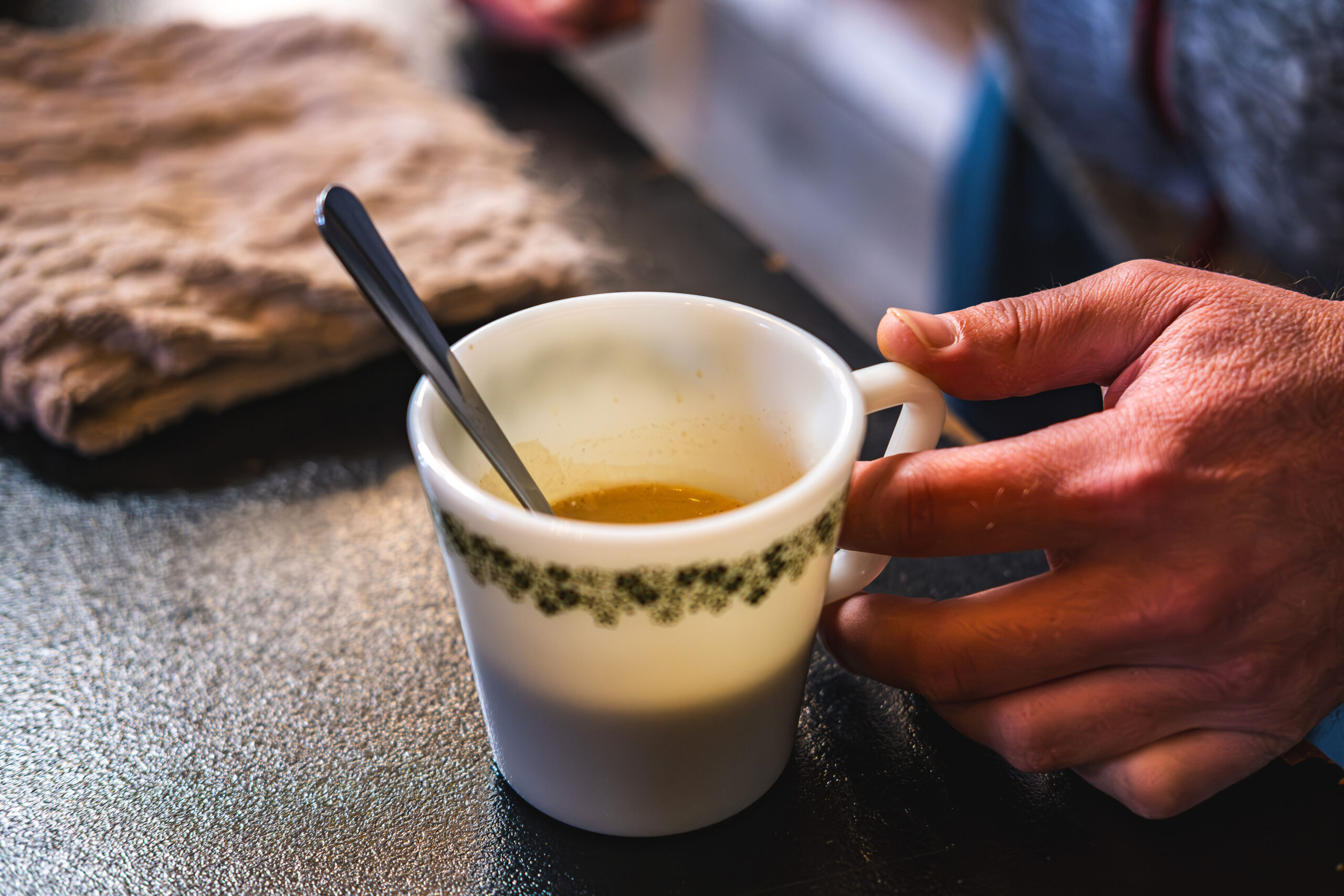 Close-up of a person's hand holding a vintage white coffee cup with a green floral pattern, resting on a dark textured surface.