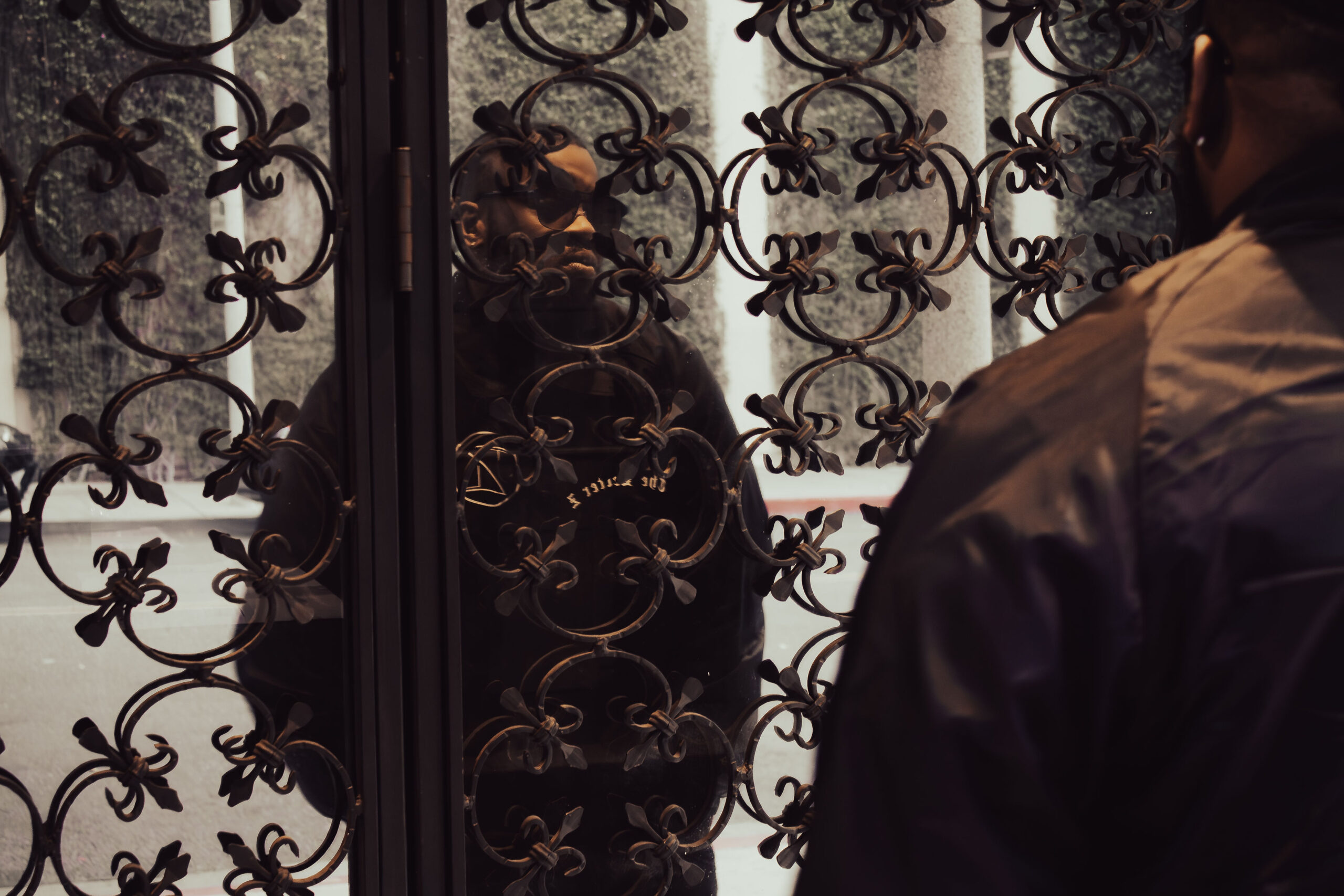 A man wearing sunglasses looking intensely through an ornate black wrought-iron gate, framed by another individual's shoulder in the foreground.
