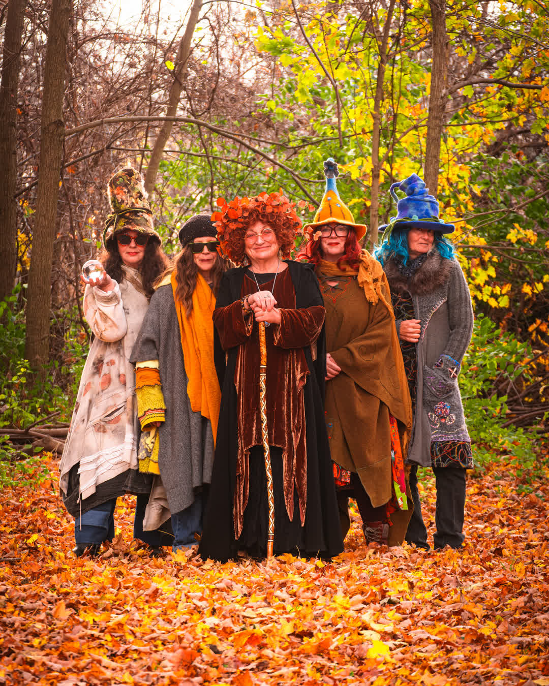 Five women dressed in elaborate, colorful woodland and witch-inspired costumes standing together in a forest covered in fallen orange autumn leaves.