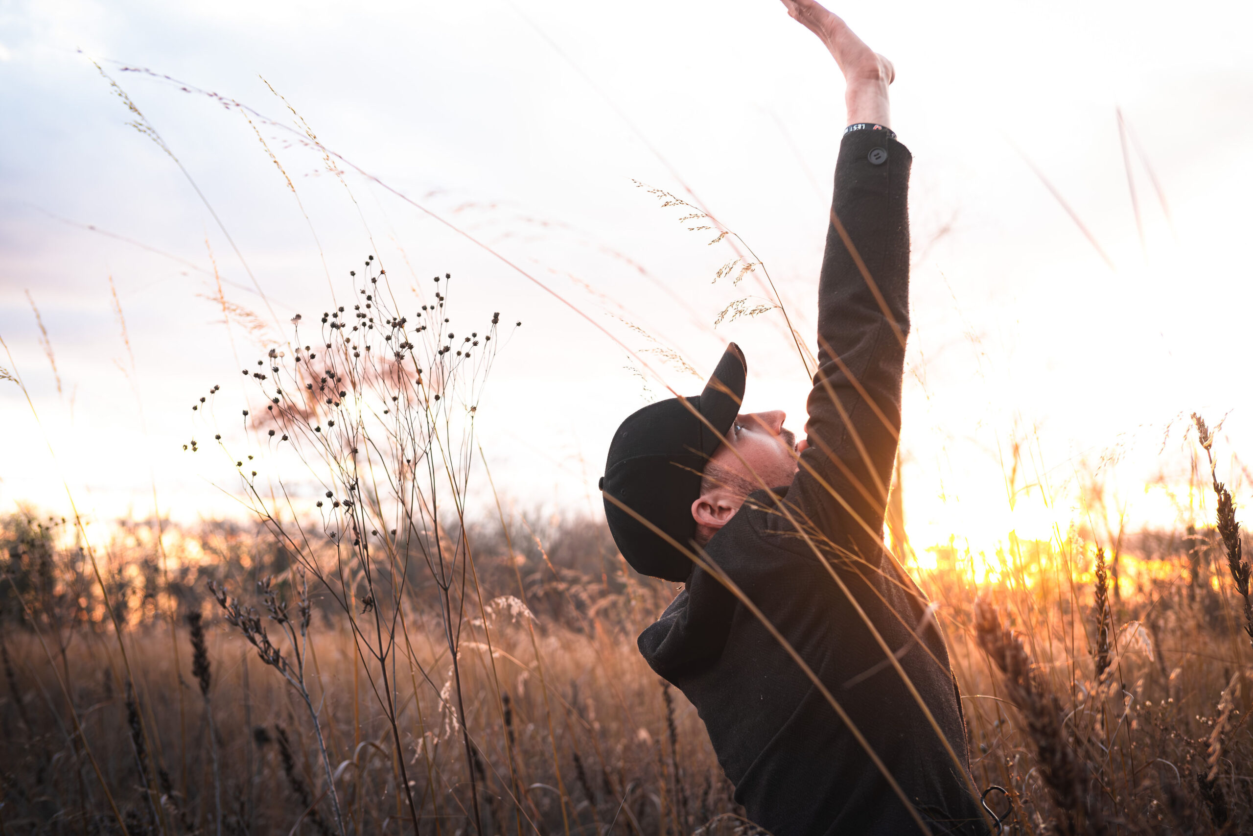 A man in a dark cap and jacket standing in a field of tall dry grass, reaching one arm upward toward the bright setting sun.