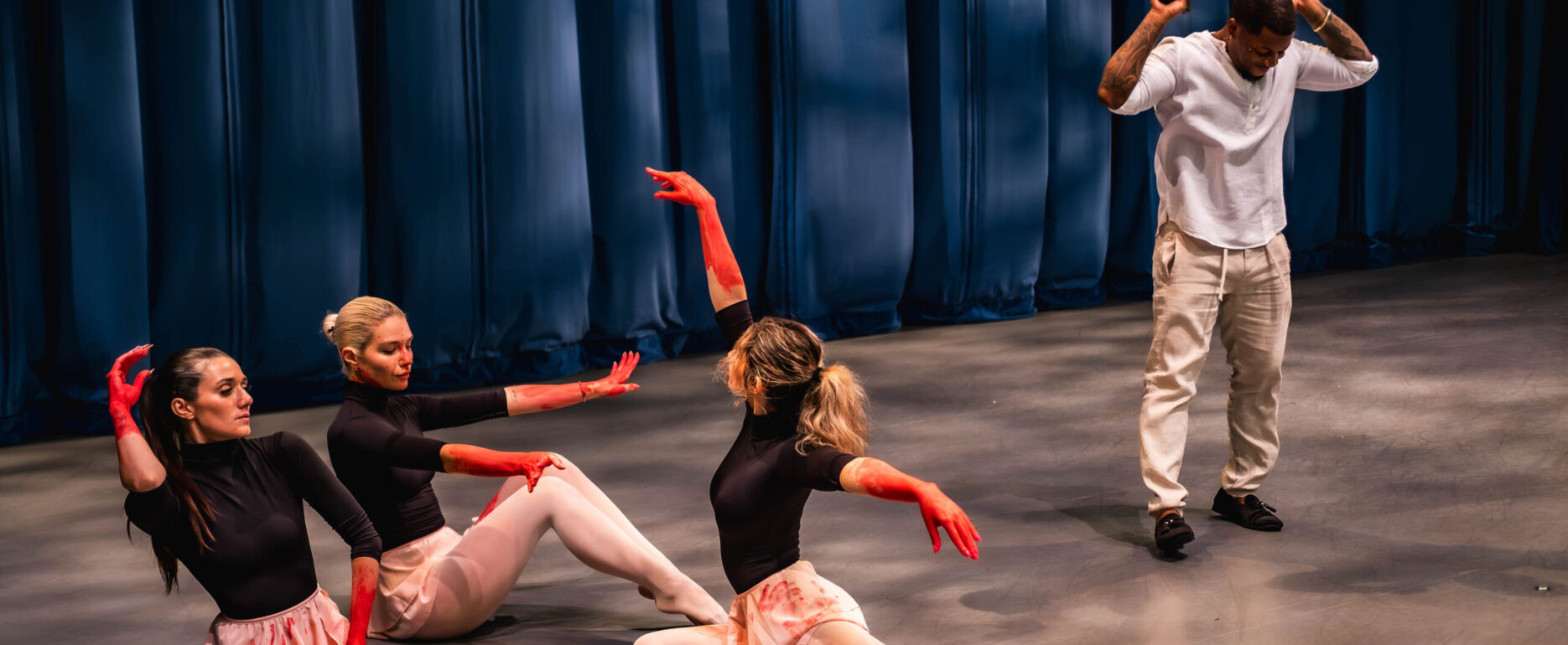 Three female dancers sitting on a stage with red paint on their hands and skirts, while a man in white clothing stands behind them making a hand gesture resembling a weapon.