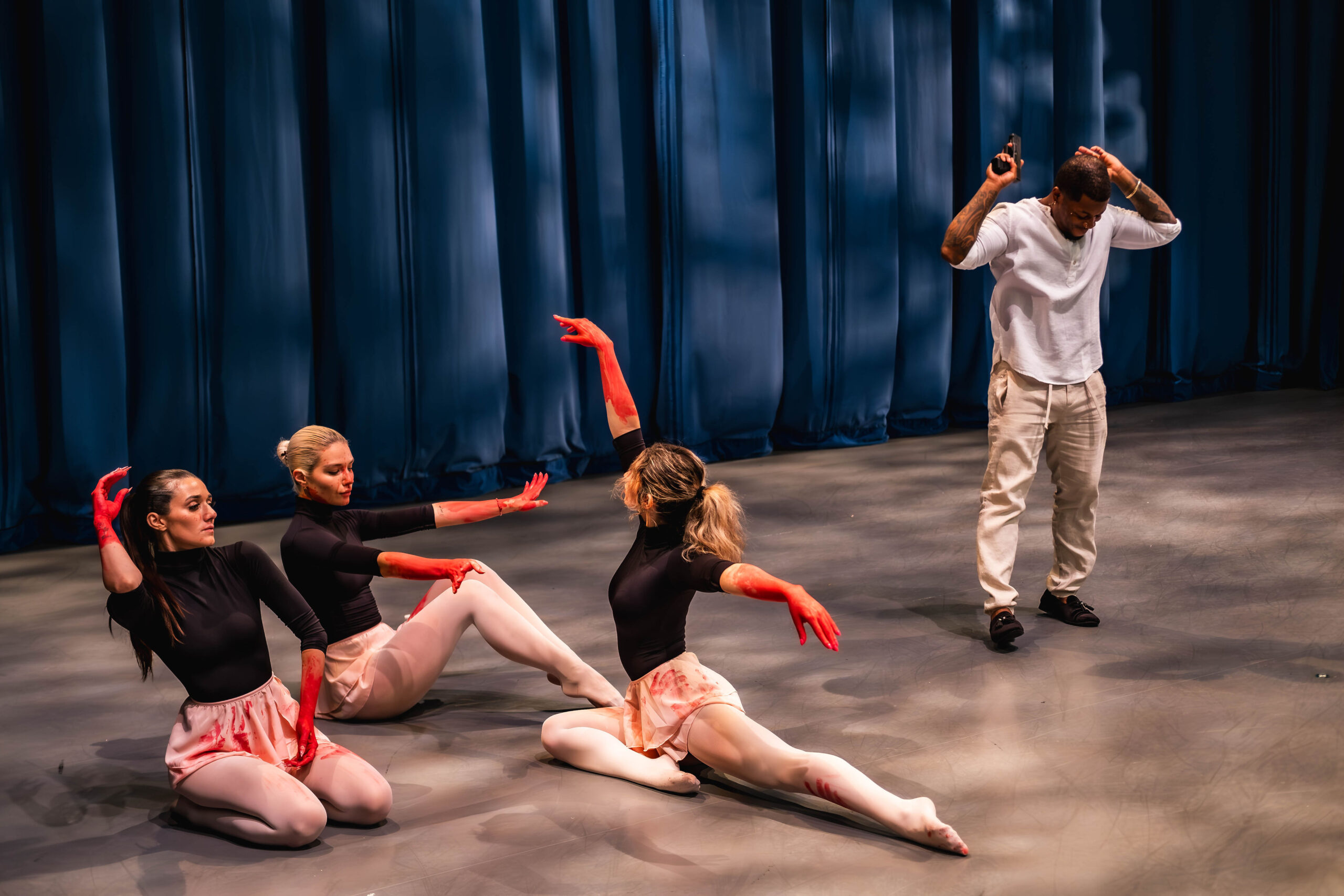 Three female dancers sitting on a stage with red paint on their hands and skirts, while a man in white clothing stands behind them making a hand gesture resembling a weapon.