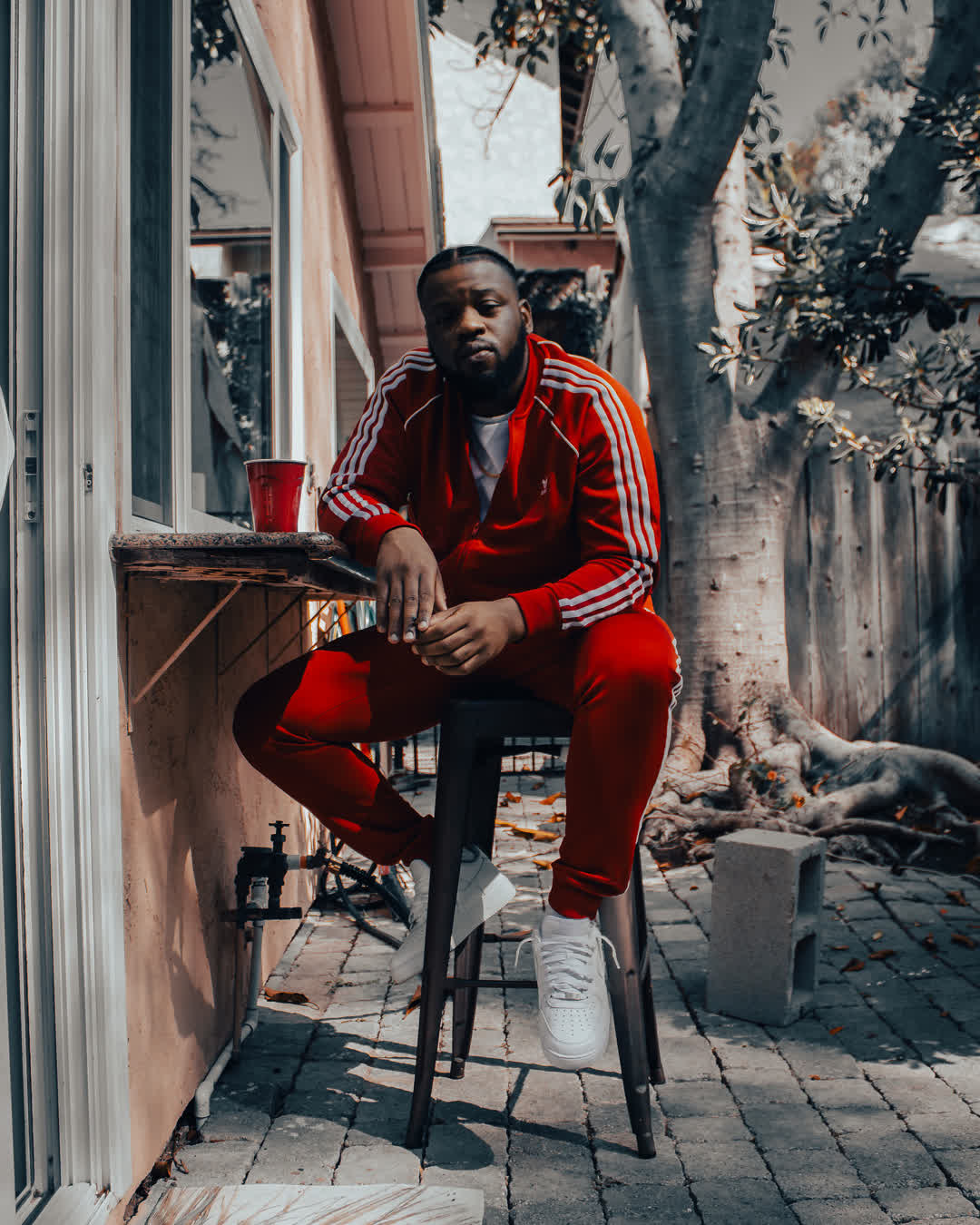 A man wearing a bright red tracksuit with white stripes sitting casually on a metal stool on a paved outdoor patio, next to a red plastic cup.