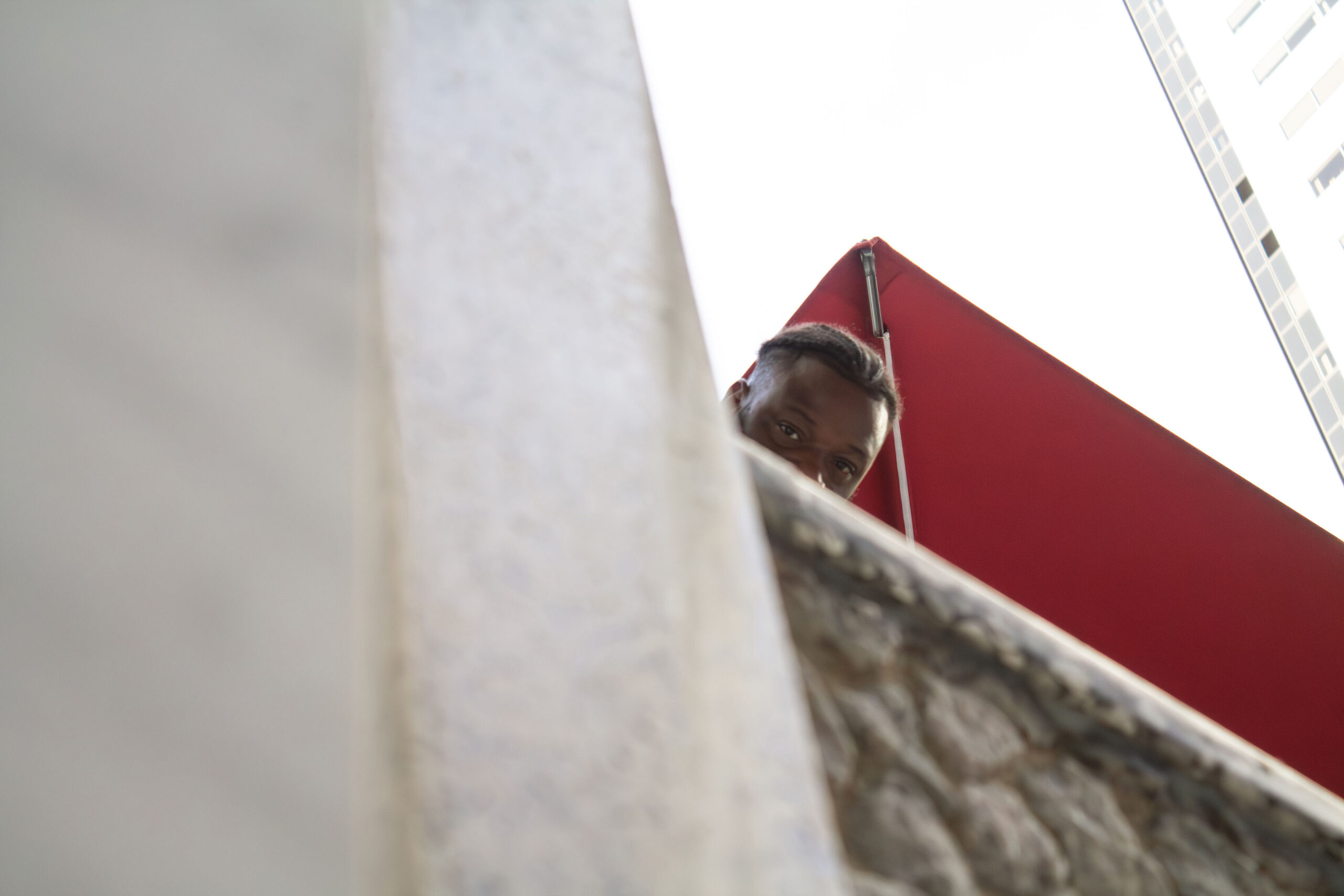 Candid street photography of a man looking down over a stone balcony wall next to a bright red awning against a bright sky.