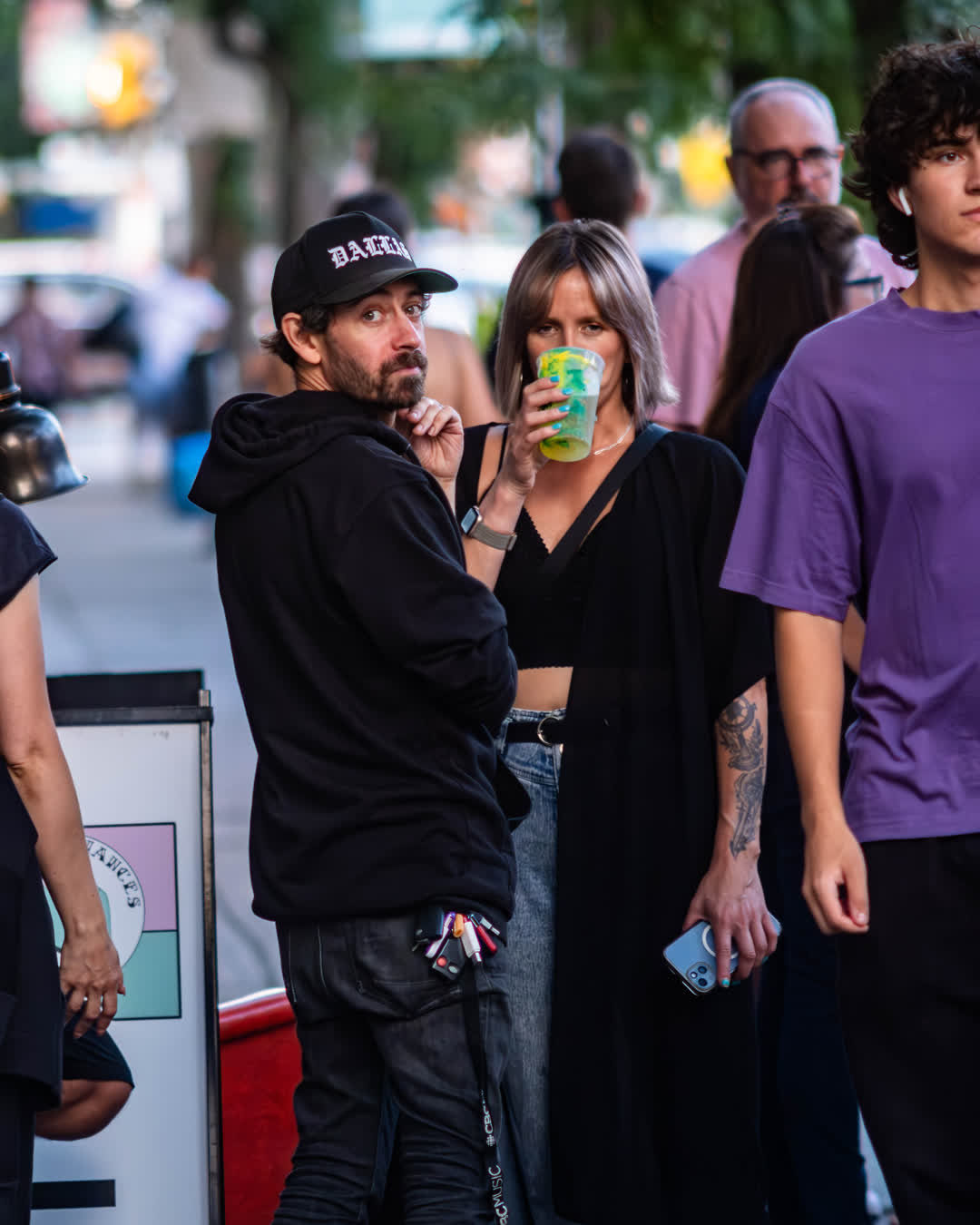 Candid street photography of a man in a black hoodie looking back at the camera while a woman next to him takes a drink from a plastic cup.