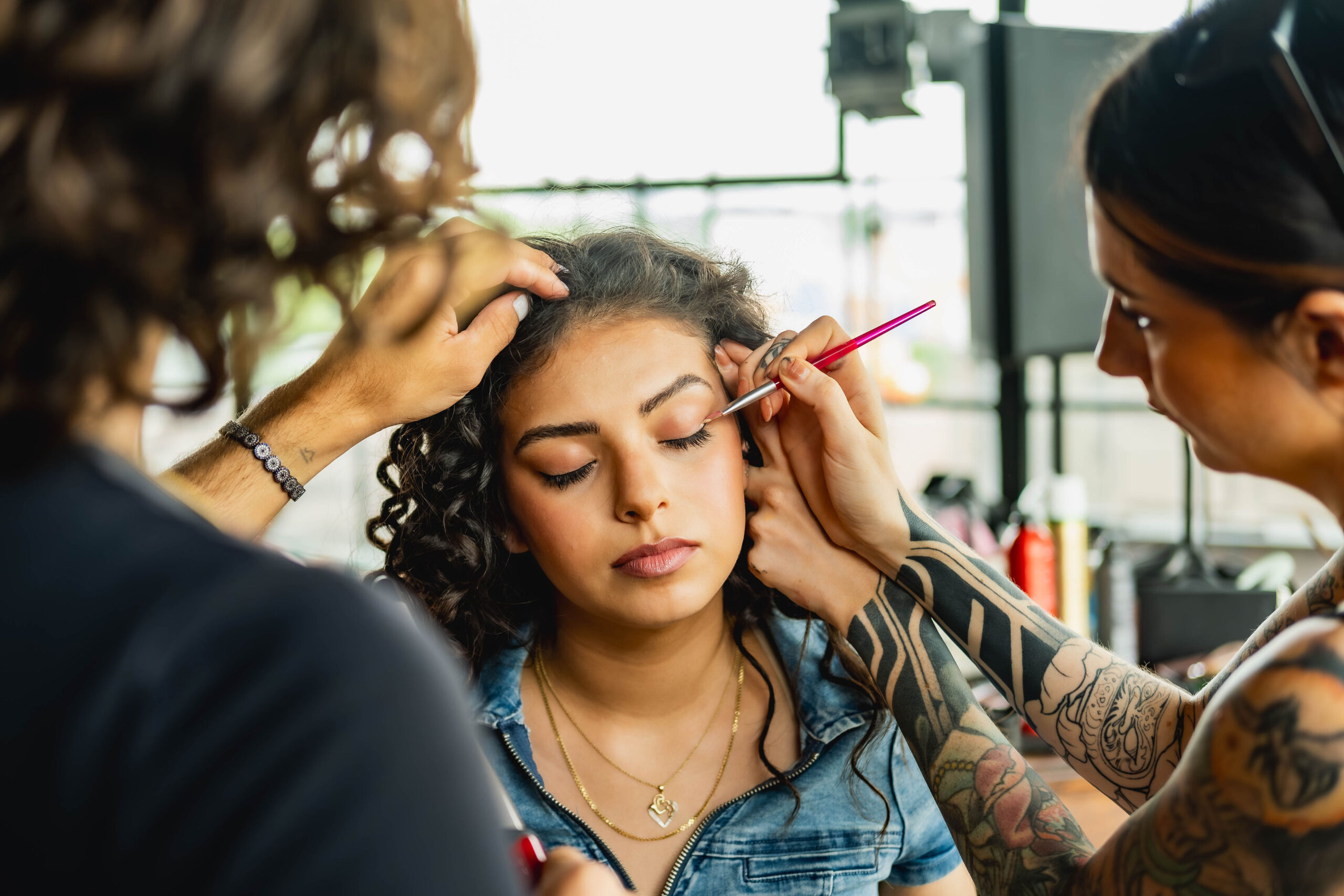 Behind the scenes photography showing two makeup artists applying eye makeup and styling a female model's hair.