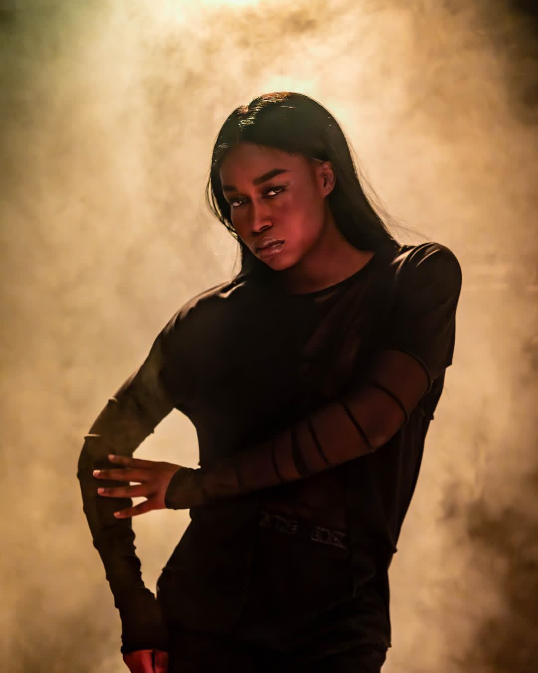 Dramatic studio portrait of a Black model wearing a black mesh top, posing powerfully against a background of warm, golden smoke.