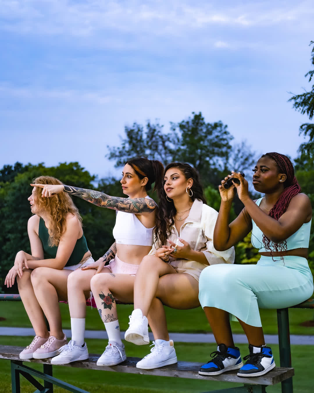 Four young women sitting together on a green park bench, looking into the distance, with one pointing forward and another holding binoculars.