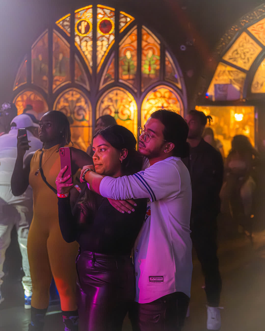 A young couple embracing and taking a selfie, illuminated by purple and orange neon lights against a backdrop of ornate stained-glass windows.