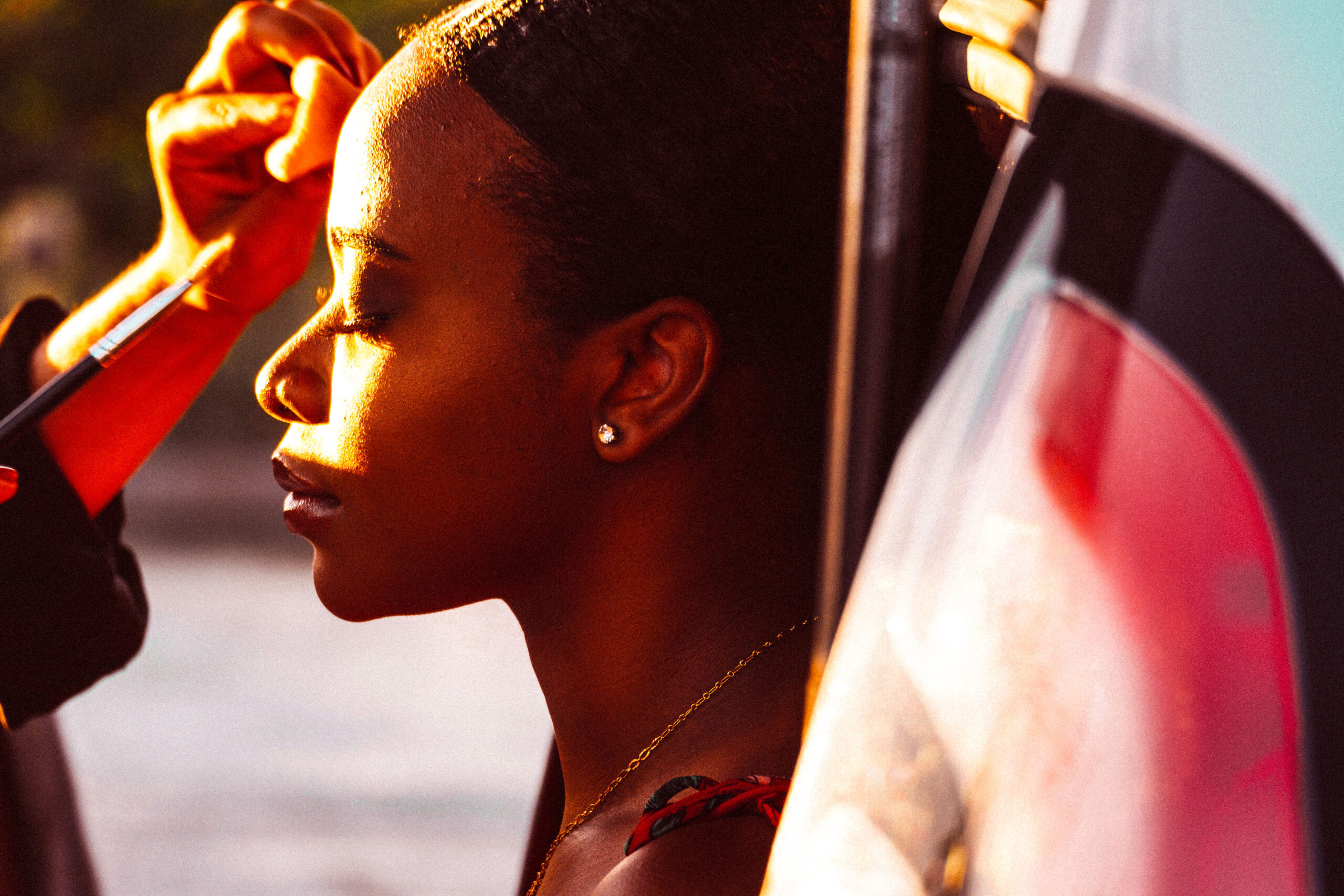 Close-up profile of a beautiful woman with her eyes closed, receiving a makeup touch-up under warm, natural sunlight.