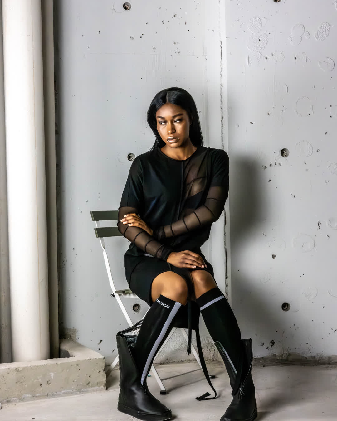 Fashion portrait of a woman in a sheer black top and knee-high boots, sitting on a minimalist chair against an unfinished concrete wall.