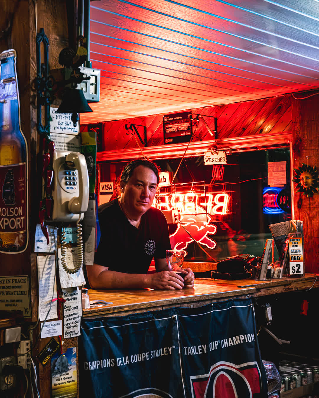 Portrait of a bartender wearing a Steeltown Garage shirt, leaning thoughtfully against a wooden bar illuminated by red neon signs.