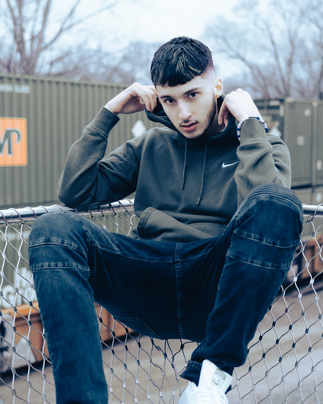 A young man in a green Nike hoodie sitting casually on a chain-link fence, set against a moody urban background with shipping containers.