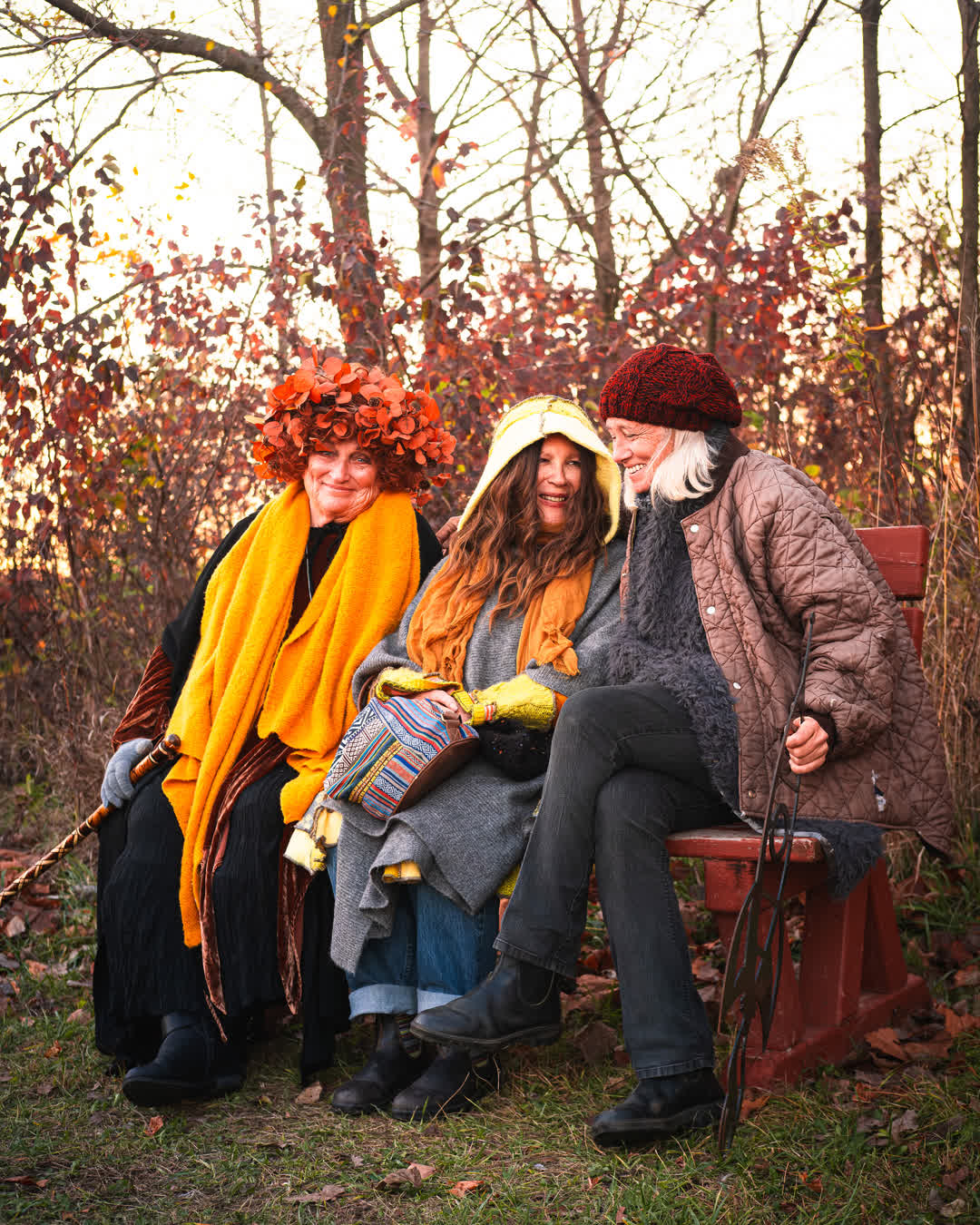 Three older women wearing eclectic, bohemian fall fashion, including a crown of leaves, laughing joyously together on a park bench.