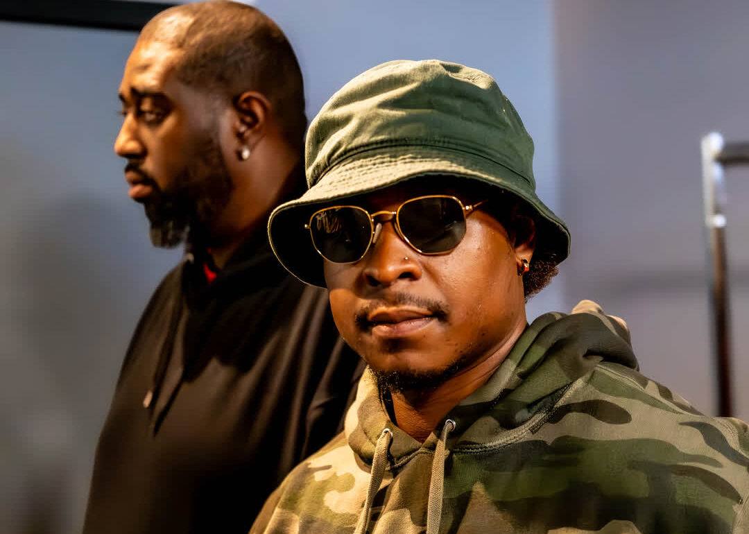 Urban portrait of a man wearing a camo hoodie, bucket hat, and dark sunglasses, with a second figure slightly out of focus in the background.