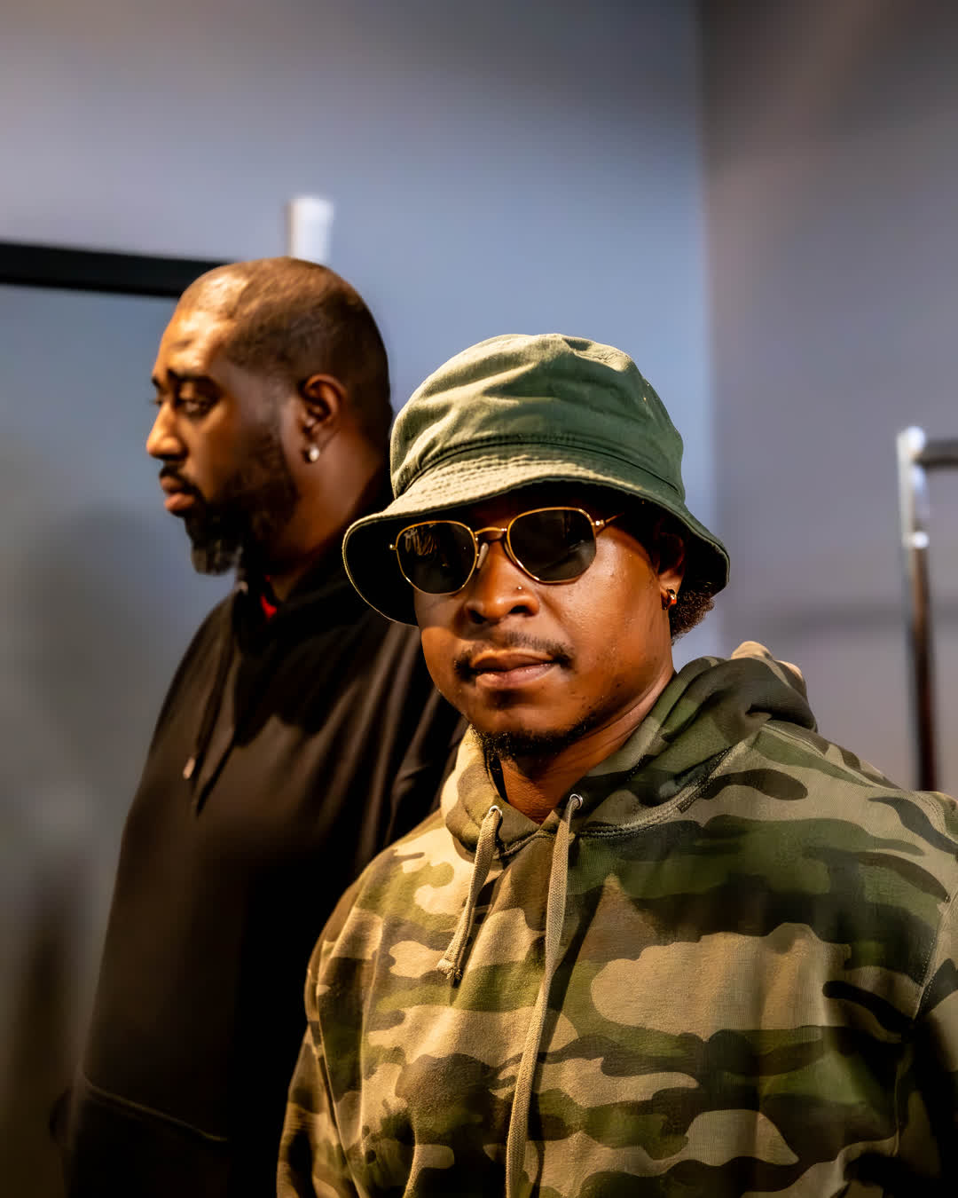 Urban portrait of a man wearing a camo hoodie, bucket hat, and dark sunglasses, with a second figure slightly out of focus in the background.