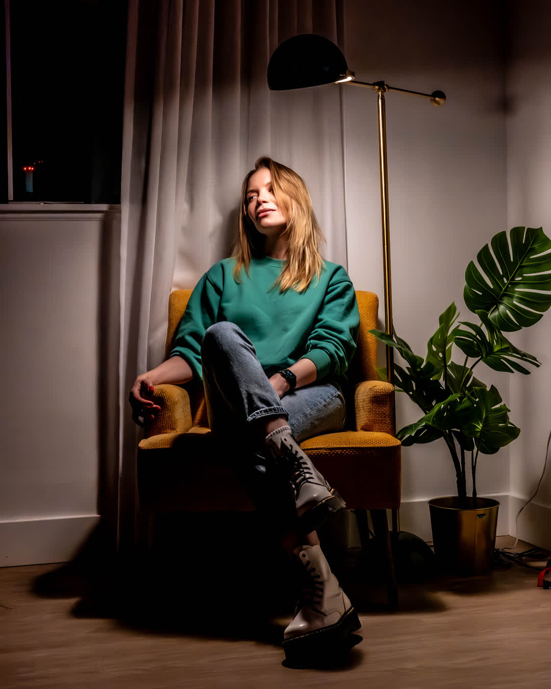 A woman wearing a green sweatshirt and boots relaxing comfortably in a mustard yellow armchair beside a modern floor lamp and a monstera plant.