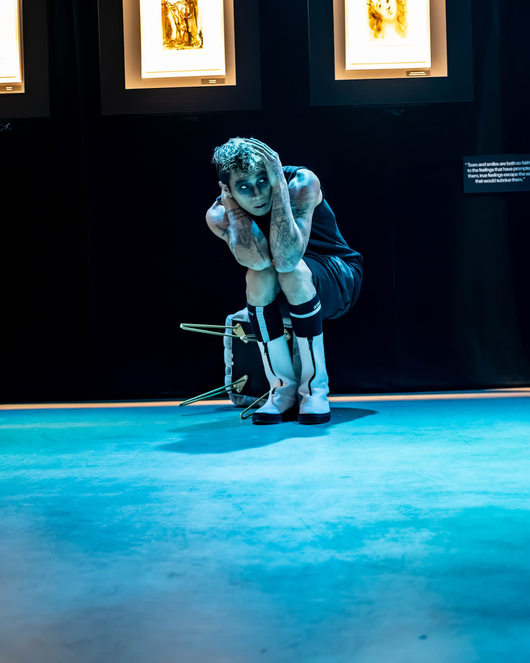 Theatrical performer with dramatic makeup crouching on an overturned chair under blue stage lighting in an art gallery.