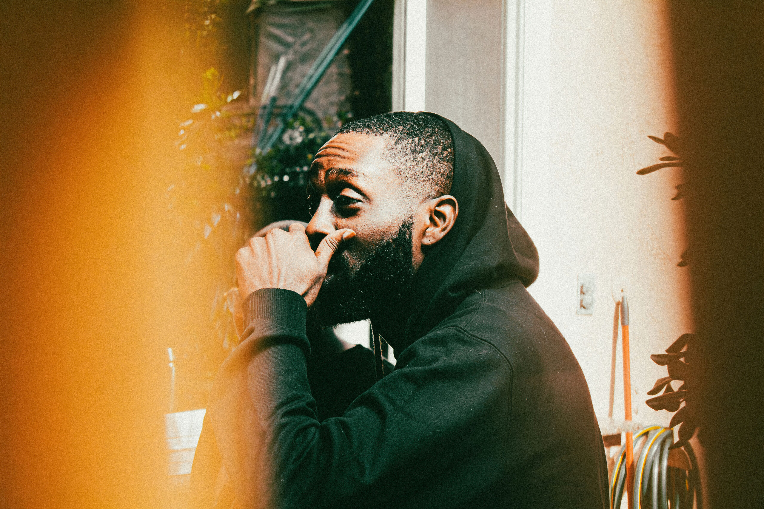 Candid portrait of a bearded man in a black hoodie looking away thoughtfully, with a warm orange light flare in the foreground.
