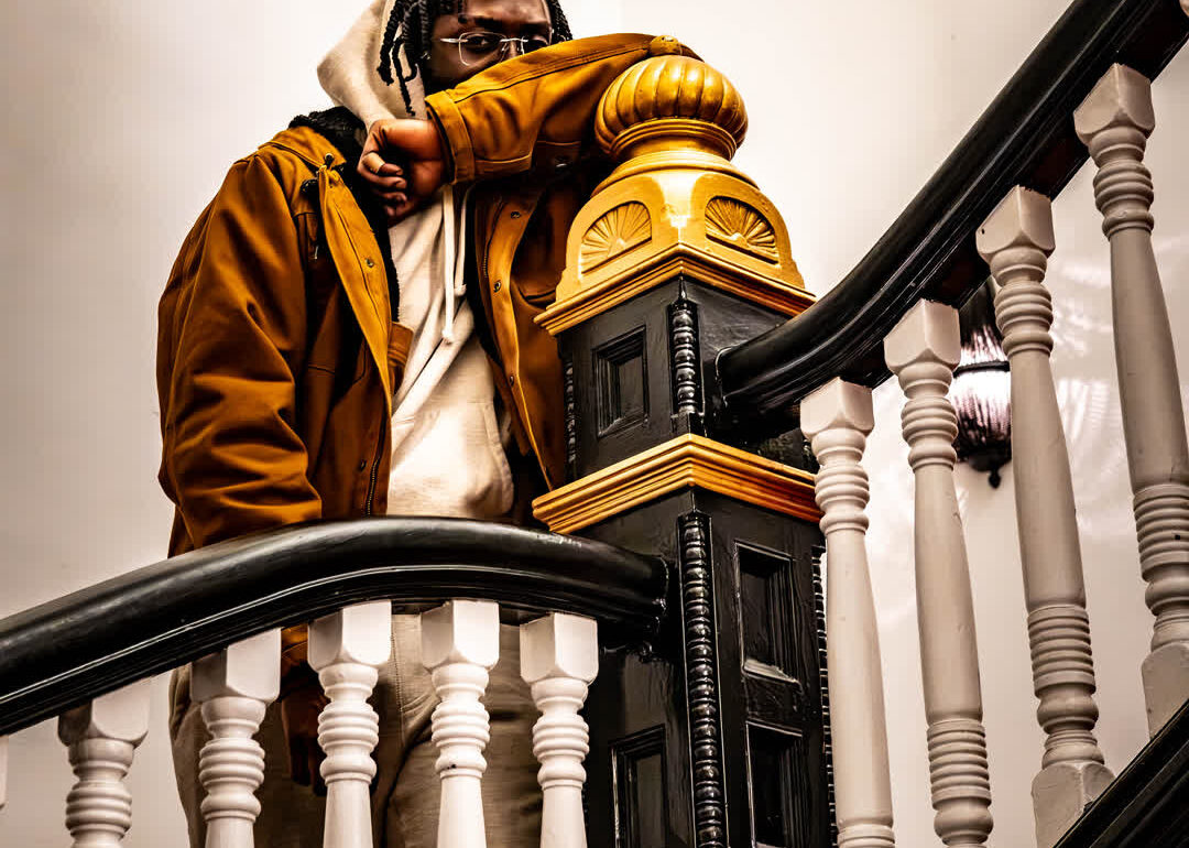 Man wearing a tan jacket and glasses leaning against a grand black and wood staircase banister in a brightly lit hallway.