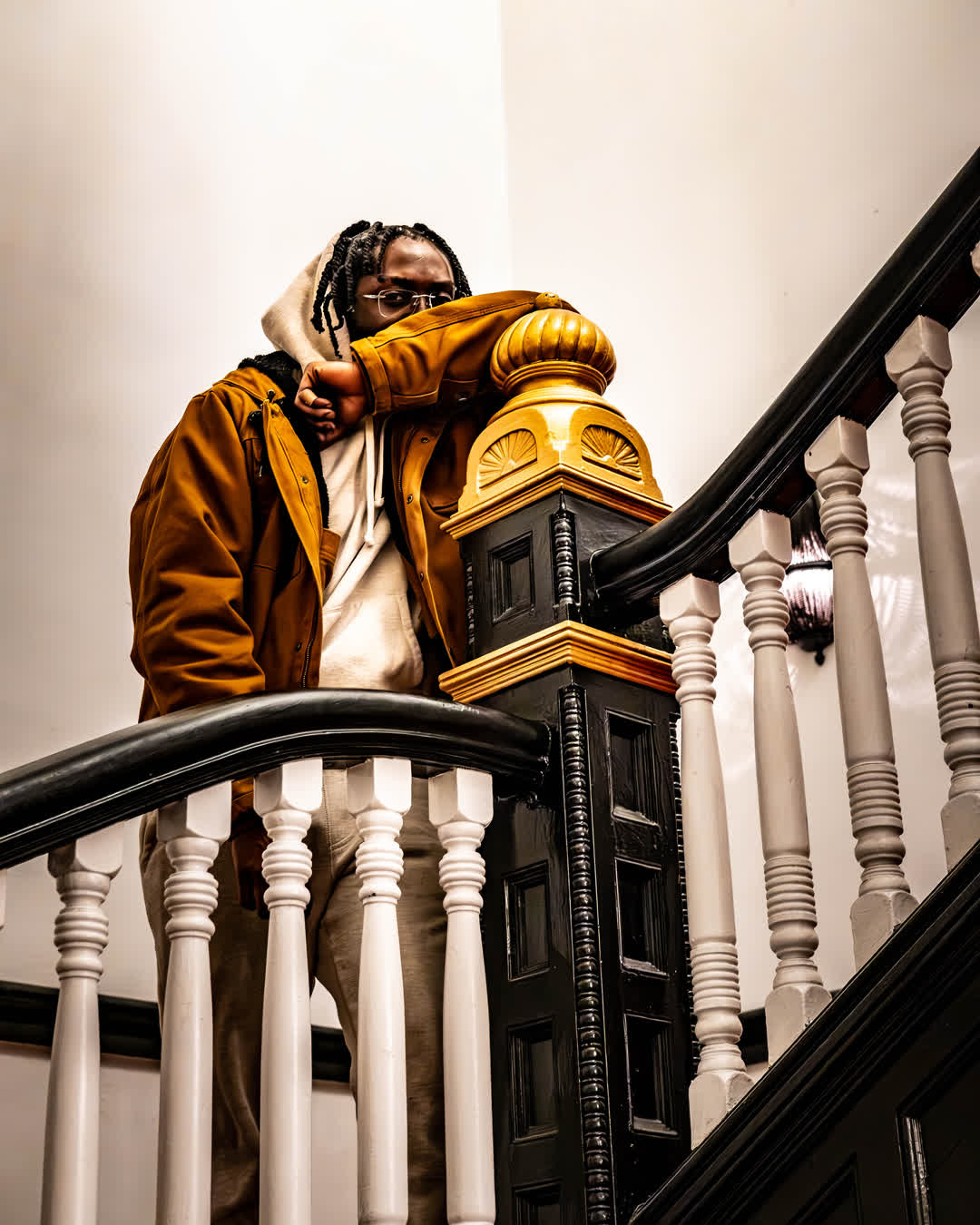 Man wearing a tan jacket and glasses leaning against a grand black and wood staircase banister in a brightly lit hallway.
