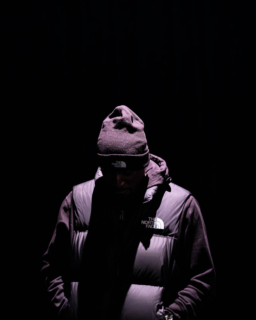 Moody portrait of a person wearing a North Face beanie and vest, illuminated from above in pitch darkness, obscuring their face in shadow.