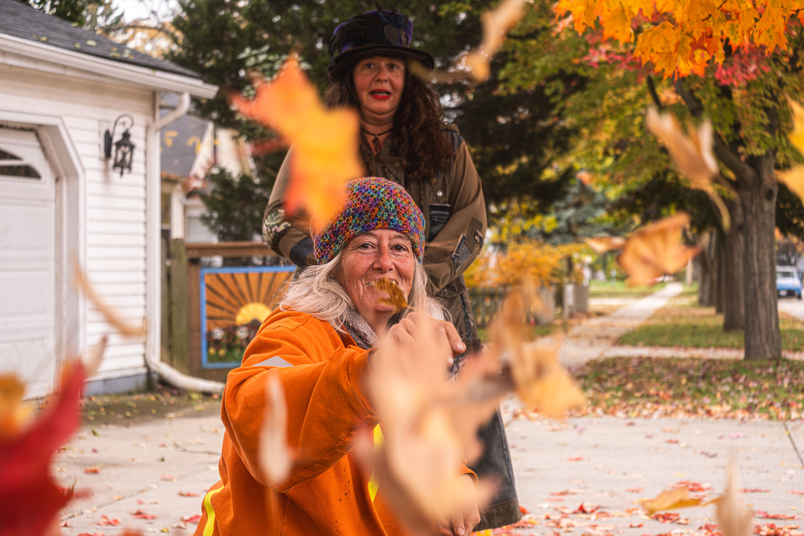 Woman in an orange jacket playfully throwing autumn leaves at the camera while another woman in a hat stands smiling behind her on a suburban sidewalk.