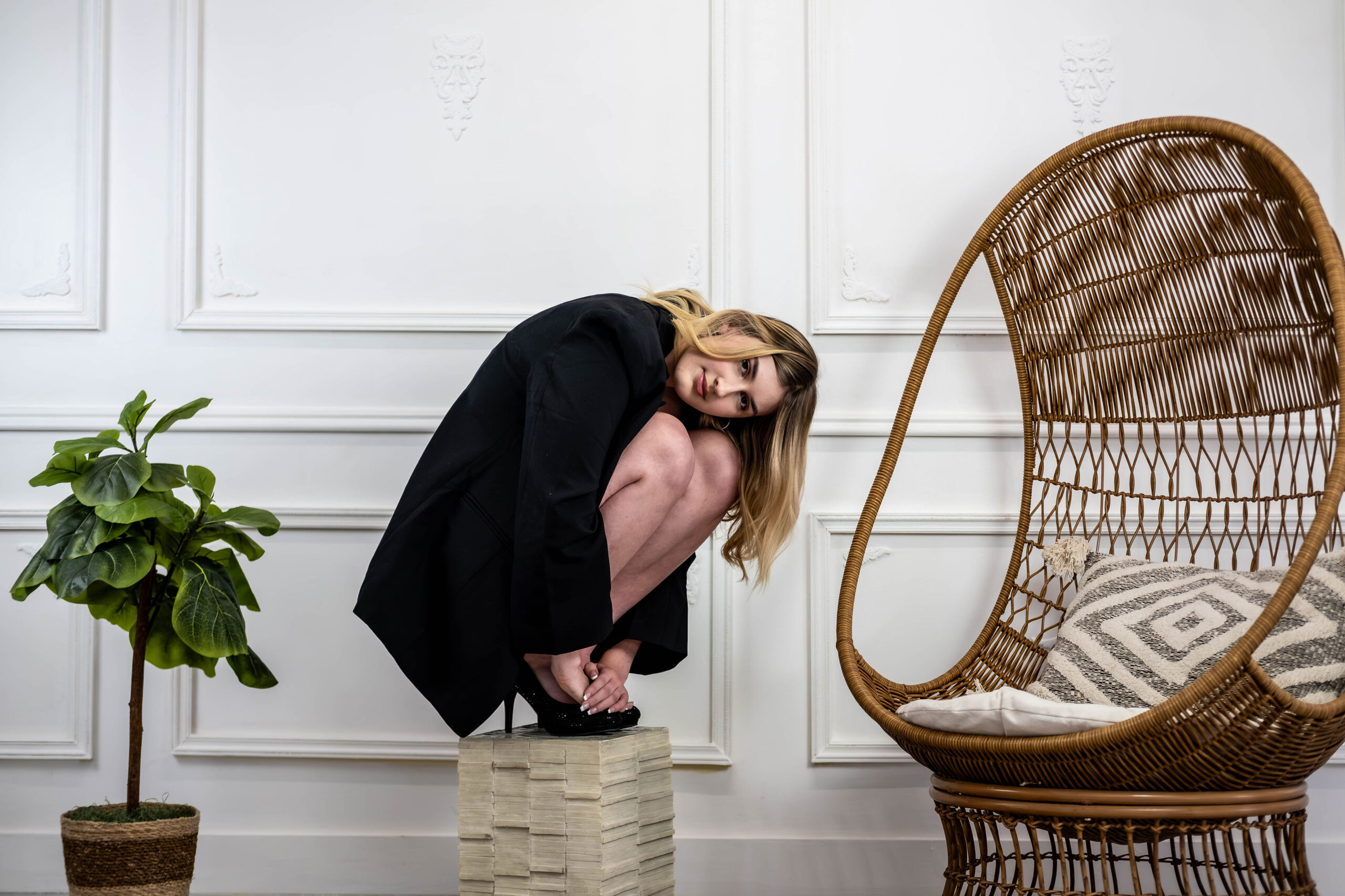 Fashion portrait of a woman in a black blazer and heels crouching on a small pedestal in a white room near a wicker chair and artificial plant.
