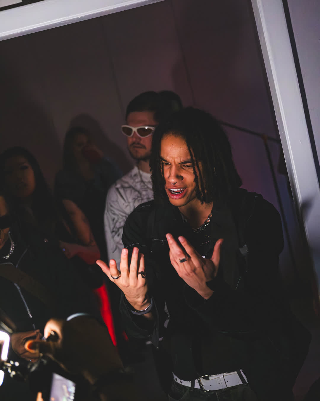 Candid event photo of a man with dreadlocks speaking passionately with his hands raised, surrounded by people in a dimly lit room with pink and purple lights.
