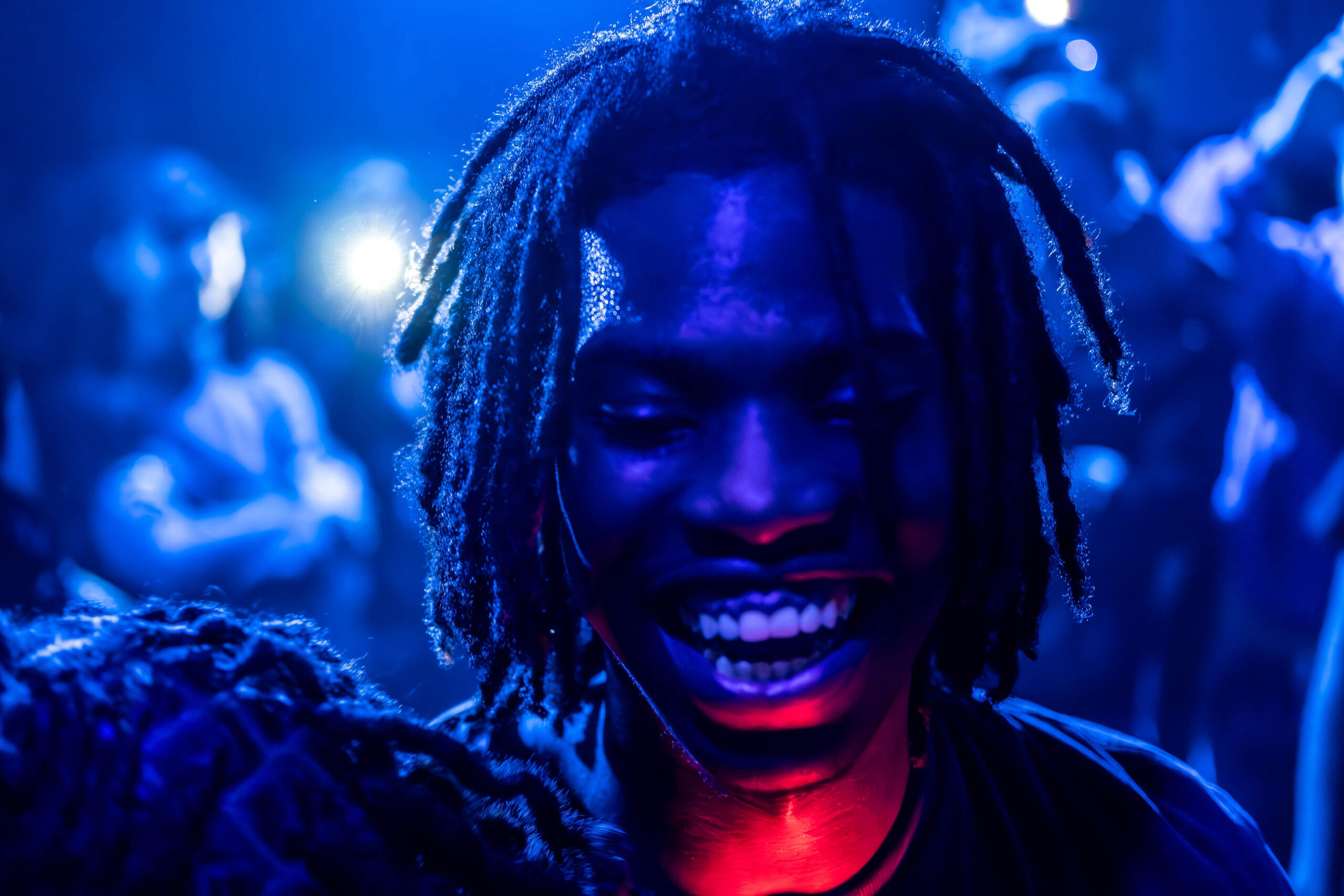 Candid portrait of a smiling man with dreadlocks illuminated by deep blue concert lighting in a crowded venue.