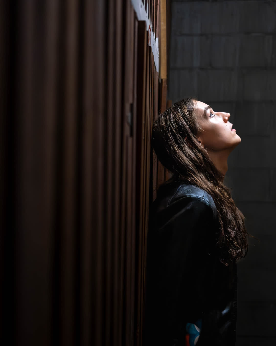 Cinematic side profile of a young woman with dark hair leaning against a textured wood wall, looking upward into a dramatic beam of light.