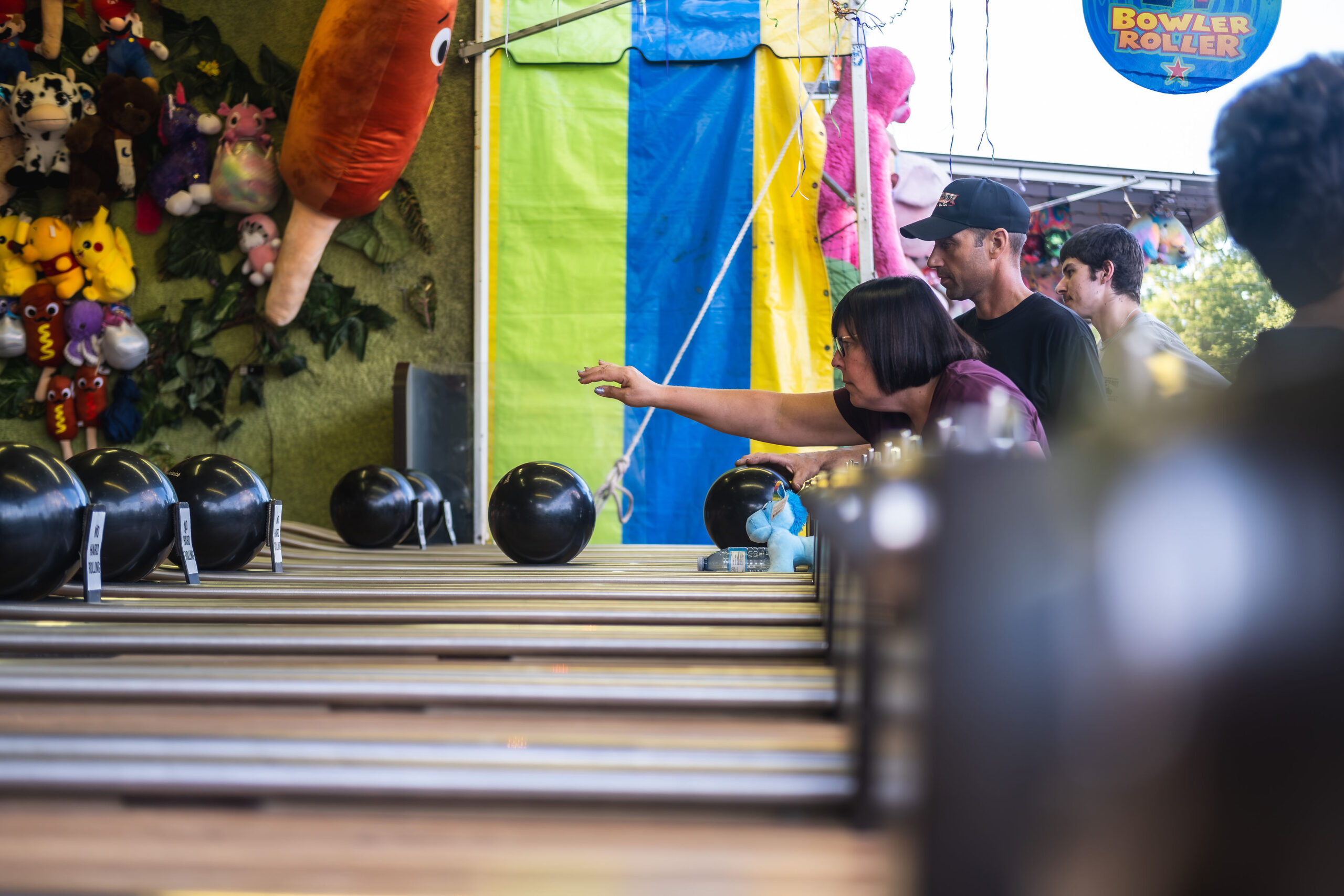 Candid photography of people playing a wooden Bowler Roller carnival game with bright plush prizes in the background.