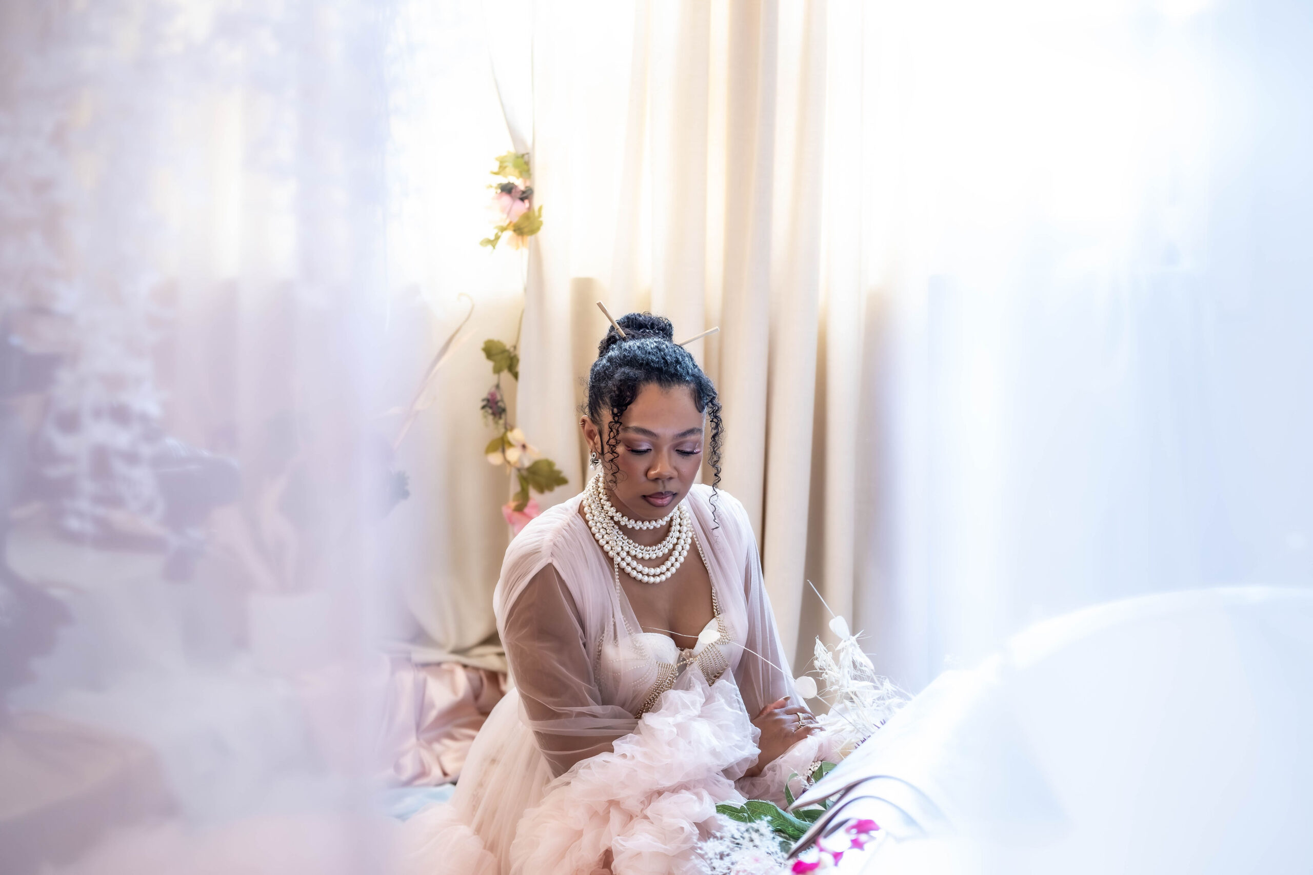 Ethereal fashion portrait of a woman wearing a ruffled pink dress and layered pearl necklaces, sitting peacefully by a brightly lit window.